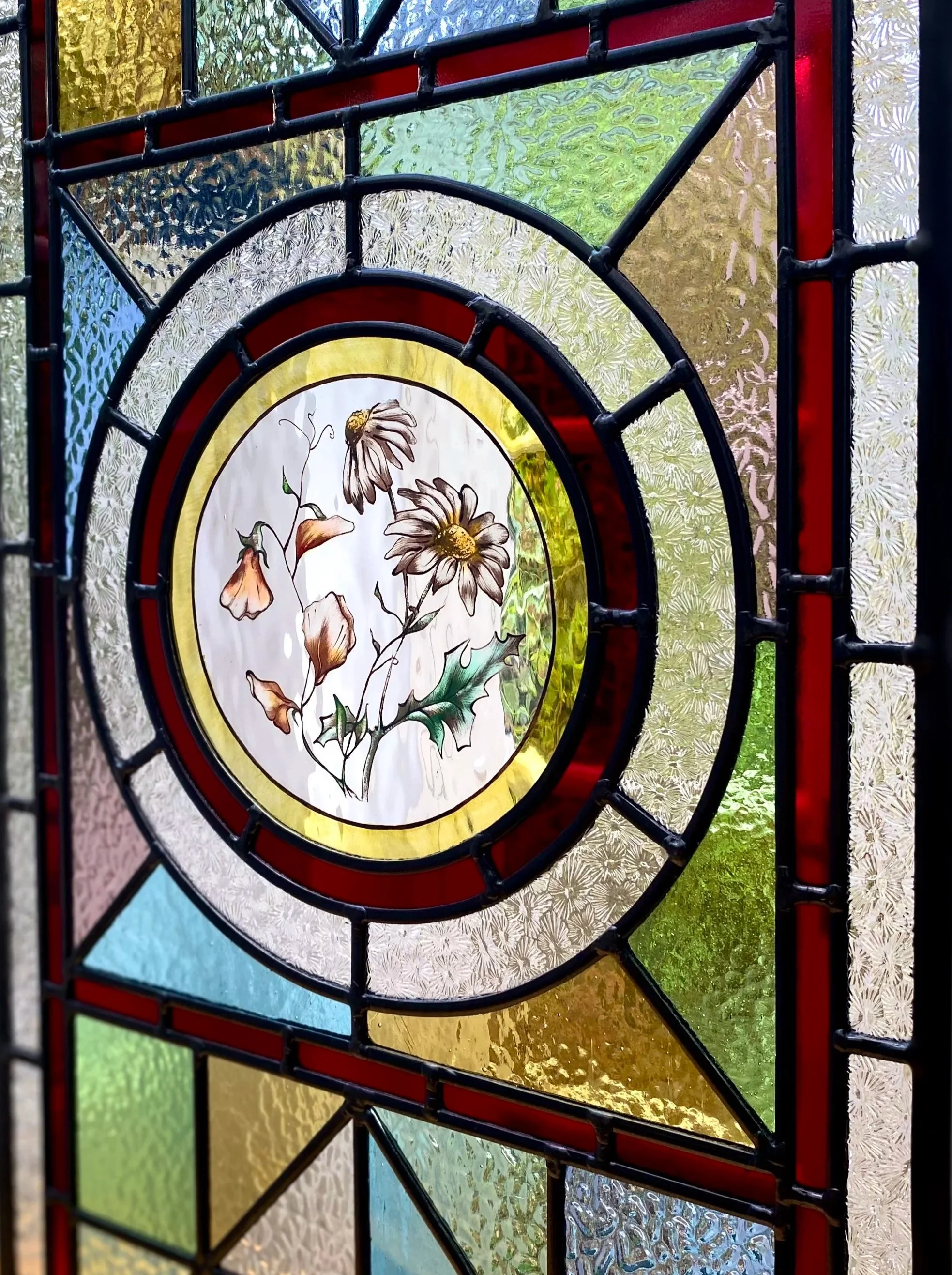 Colorful stained glass window with a central floral design featuring daisies and holly leaves, surrounded by geometric sections in various colors.