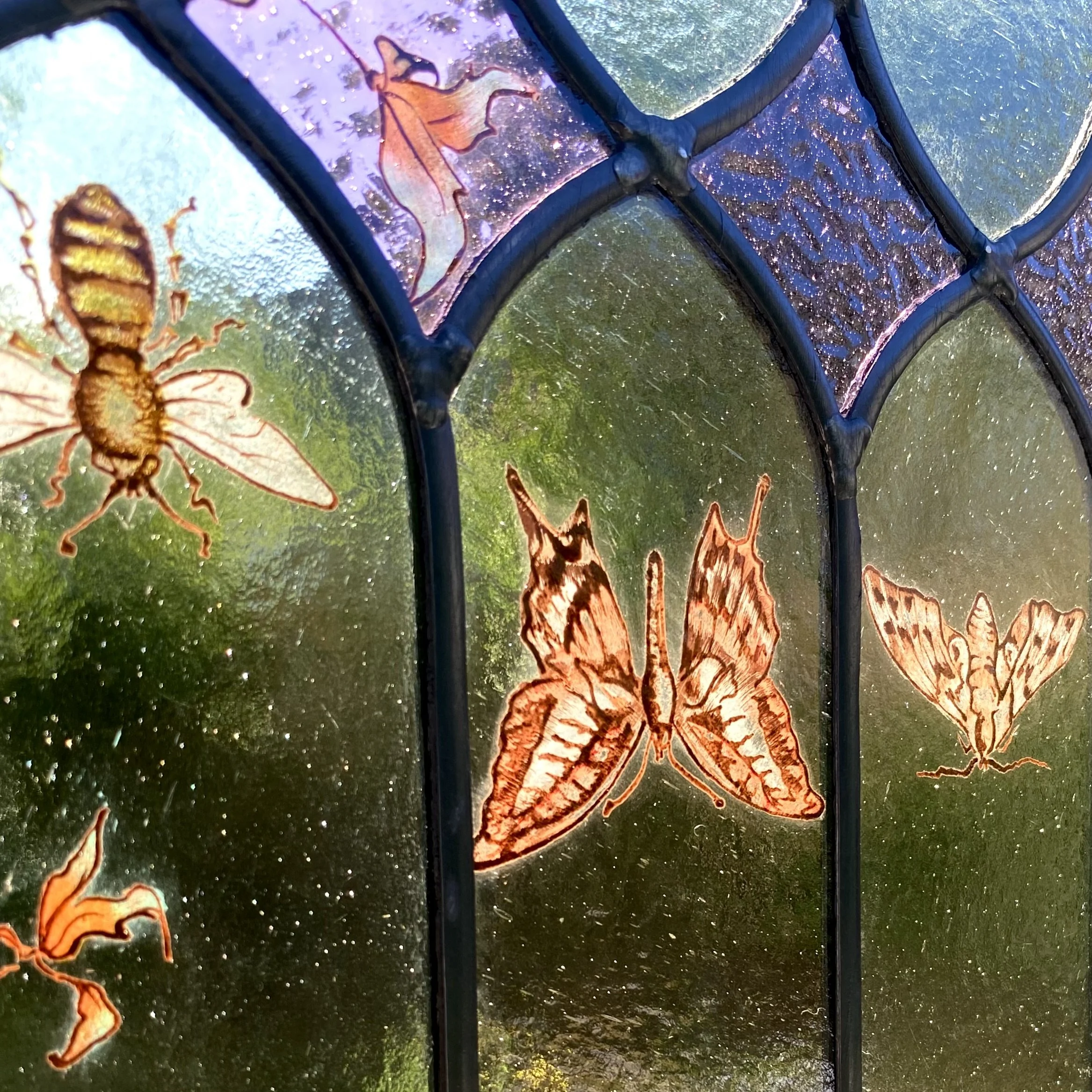 Close-up of a stained glass window featuring orange butterfly and bee designs, with water droplets on the glass and greenery outside.