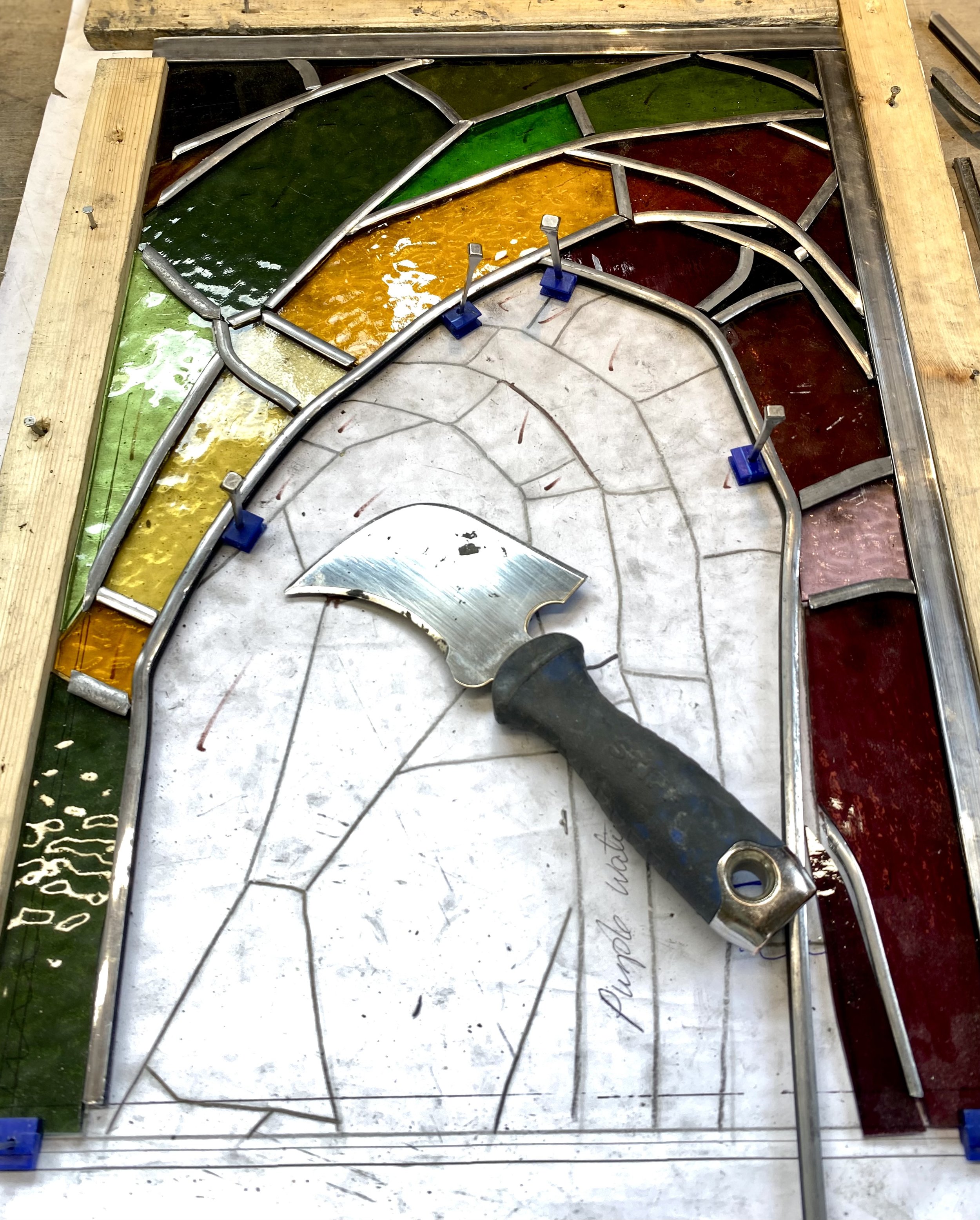 Colorful stained glass window panel, with sections in green, yellow, red, and brown, in a metal frame, laying on a worktable with wooden borders and tools.
