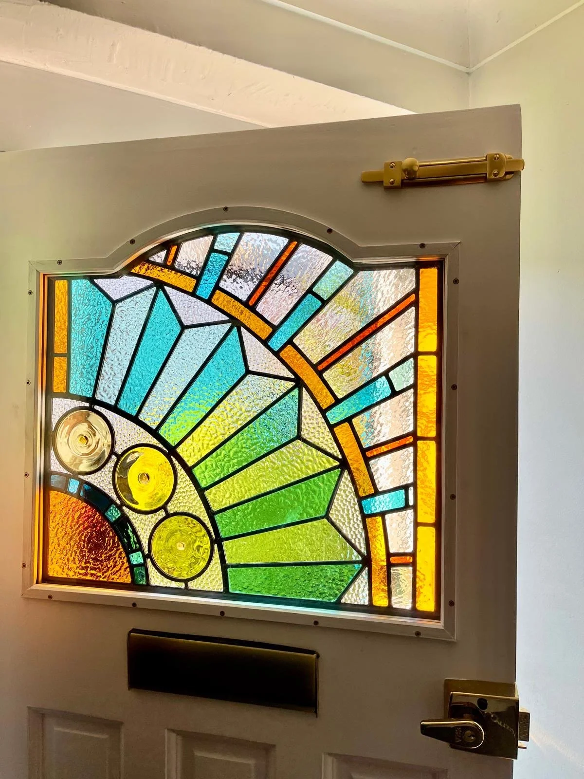 Colorful stained glass window with geometric shapes, featuring shades of blue, green, yellow, orange, and red.  Commissioned for South East London Brockley area sunburst design, designed and built by Zoe Henderson Polatch