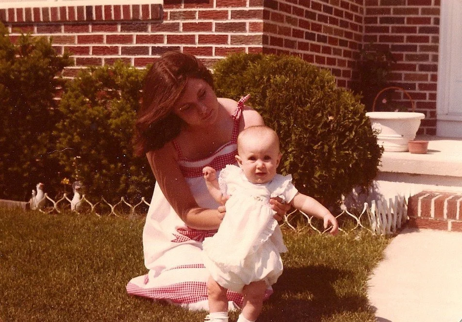 A young girl kneeling on the grass, helping a baby girl stand outdoors next to a brick house with potted plants and a white picket fence.