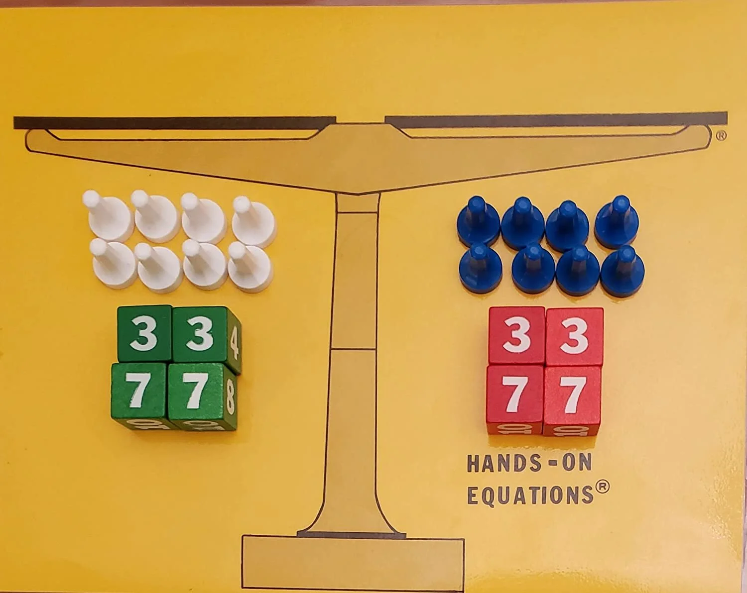 A yellow board with a black and white line drawing of a balance scale. On the left side of the scale, there are six white game pieces and three green blocks with the numbers 3 and 7. On the right side, there are six blue game pieces and three red blocks with the numbers 3 and 7. The text underneath says 'HANDS-ON EQUATIONS'.