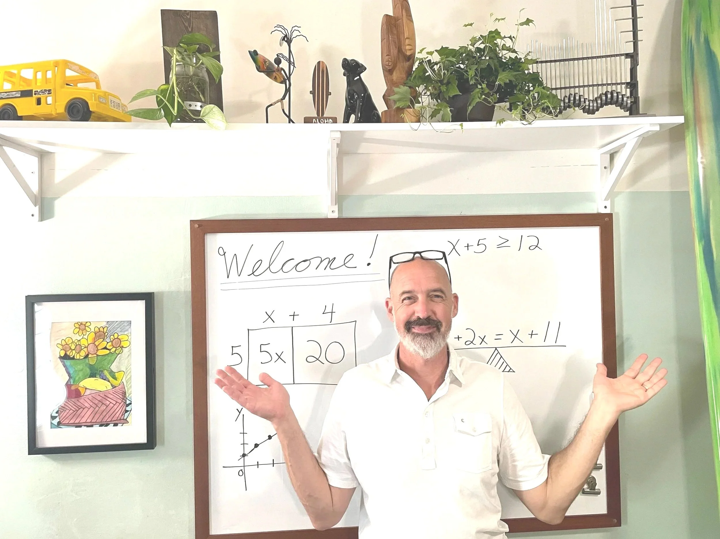 A smiling man with a beard and white shirt standing in front of a whiteboard with math equations, gesturing with open hands; classroom wall decorated with framed artwork and shelves with plants and wooden decorations.