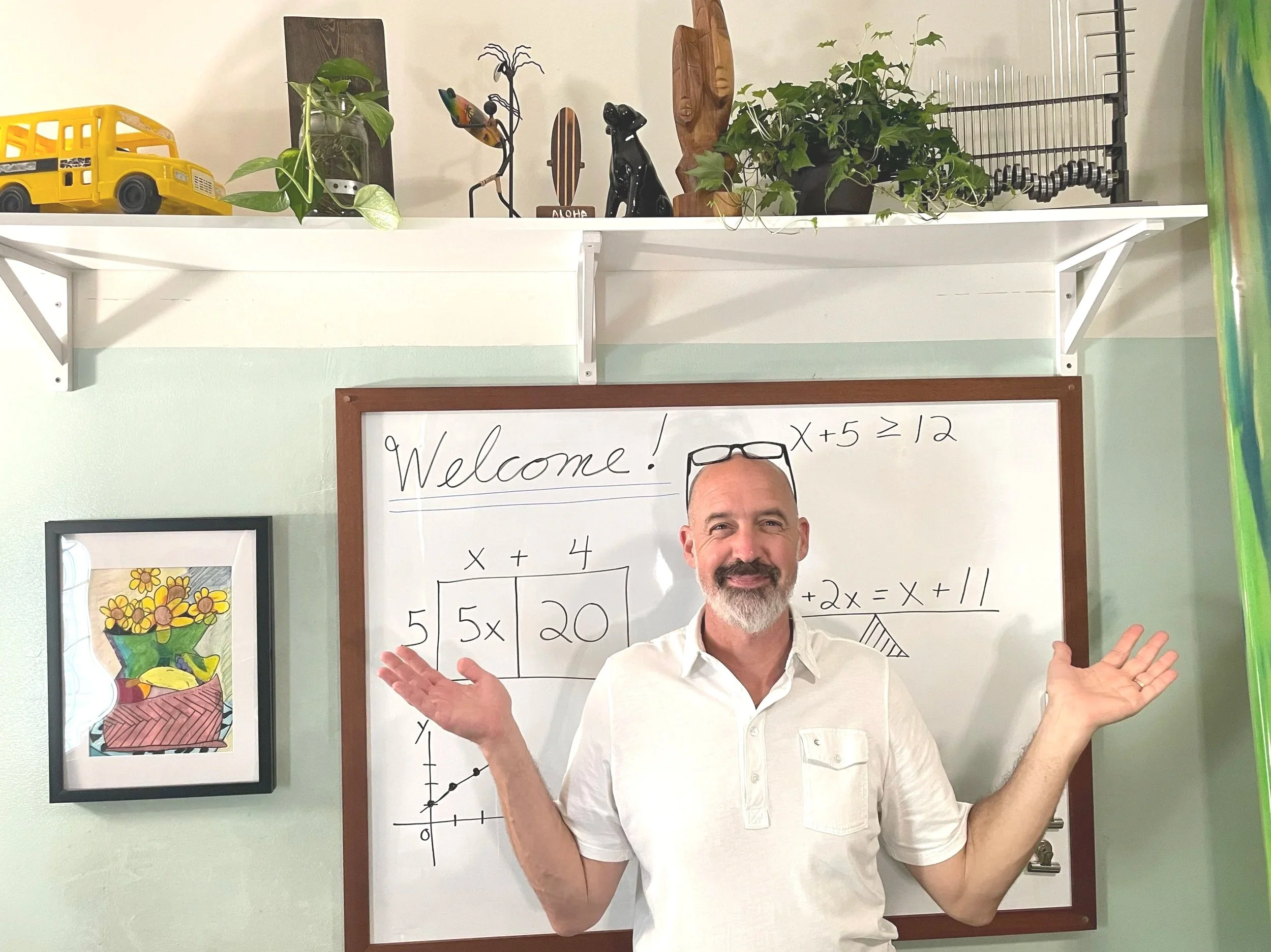 A man with a beard and mustache wearing a white shirt standing in front of a whiteboard with mathematical equations, smiling and gesturing with his hands. Behind him, there are shelves with plants and decorative wooden art pieces, and a colorful framed artwork.