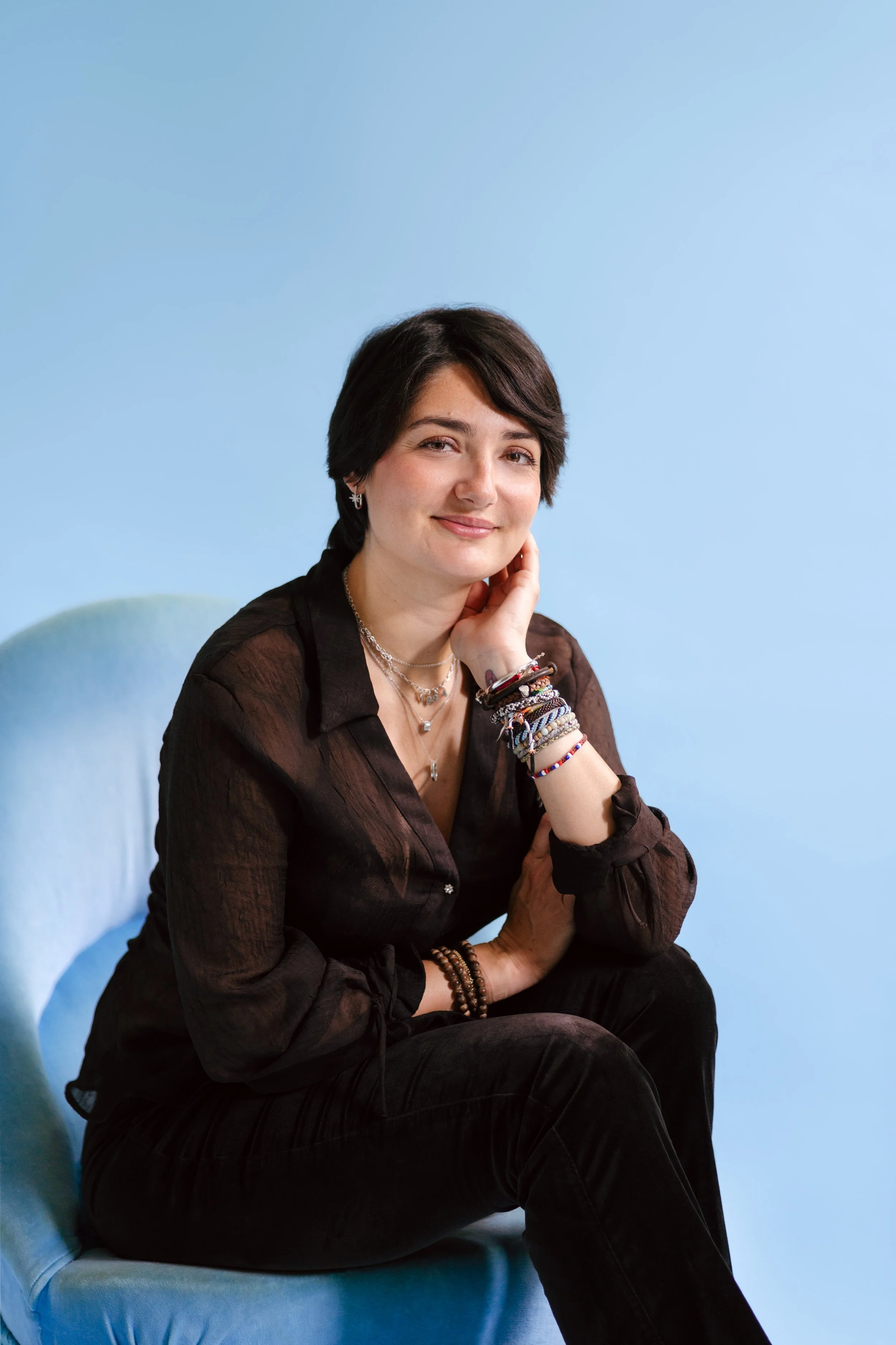 Une femme avec des cheveux courts, portant des bijoux et des bracelets colorés, assise sur un fauteuil bleu clair, souriant doucement, contre un fond bleu clair.