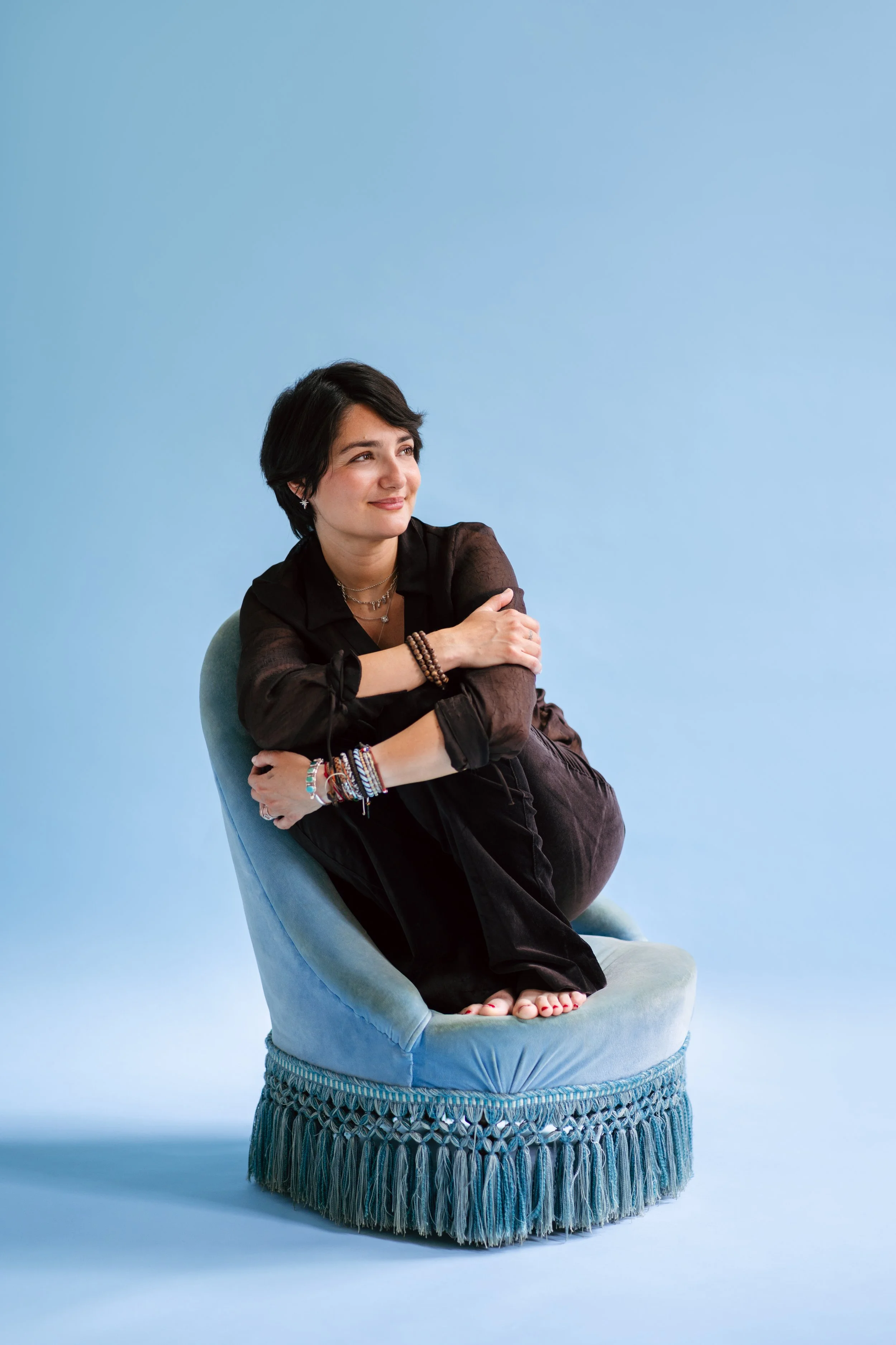 Une femme aux cheveux noirs courts, assise dans un fauteuil en velours bleu, posant de façon détendue contre le dos du fauteuil avec un fond bleu clair.