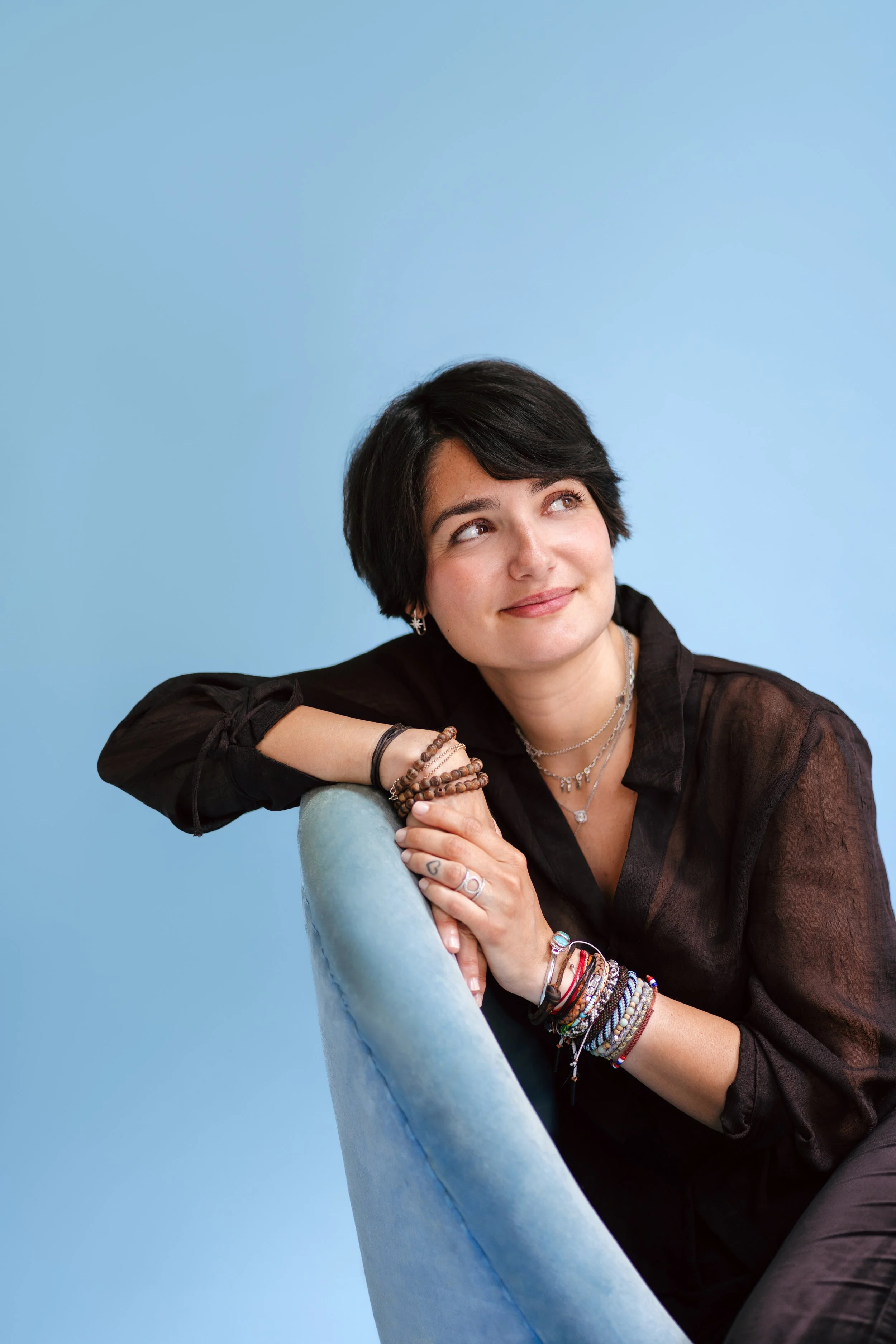 Une femme avec des cheveux noirs court, portant plusieurs colliers et bracelets, souriant et regardant vers la droite, posée contre un dossier de fauteuil beige clair avec un fond bleu clair.