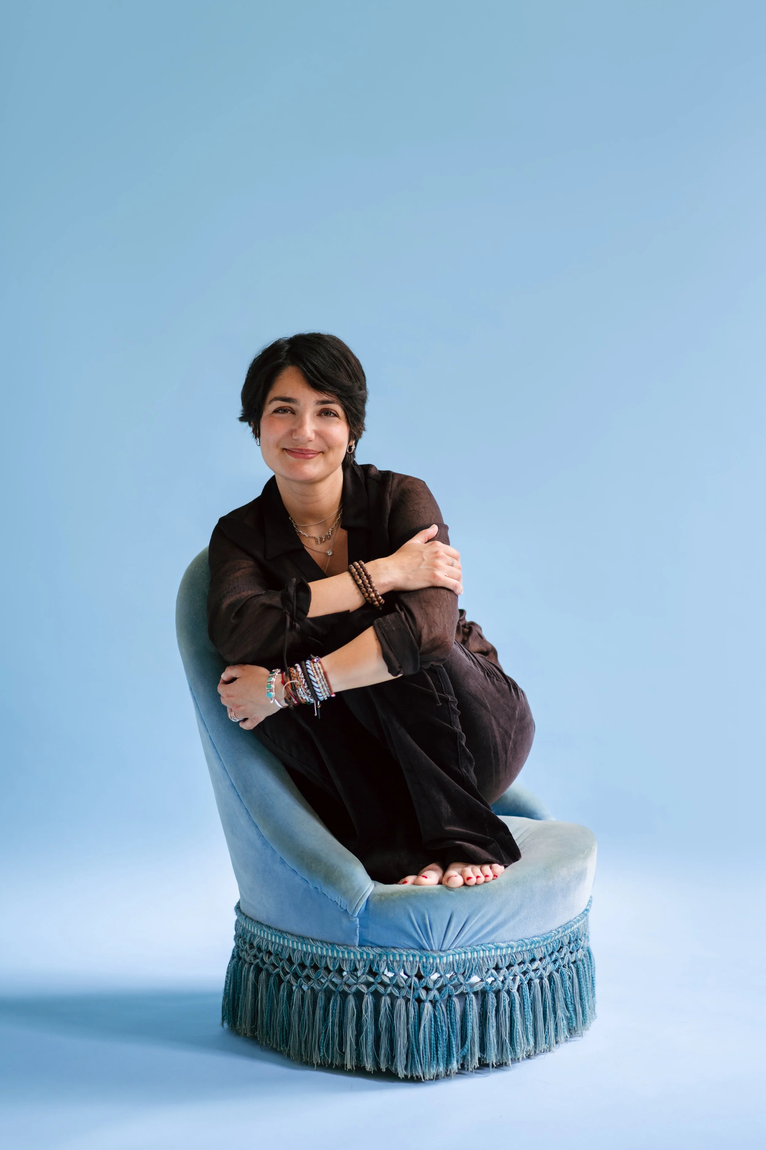 Une femme avec des cheveux courts, portant des bijoux colorés et une chemise noire, assise de manière décontractée sur un fauteuil bleu, avec un fond bleu clair.