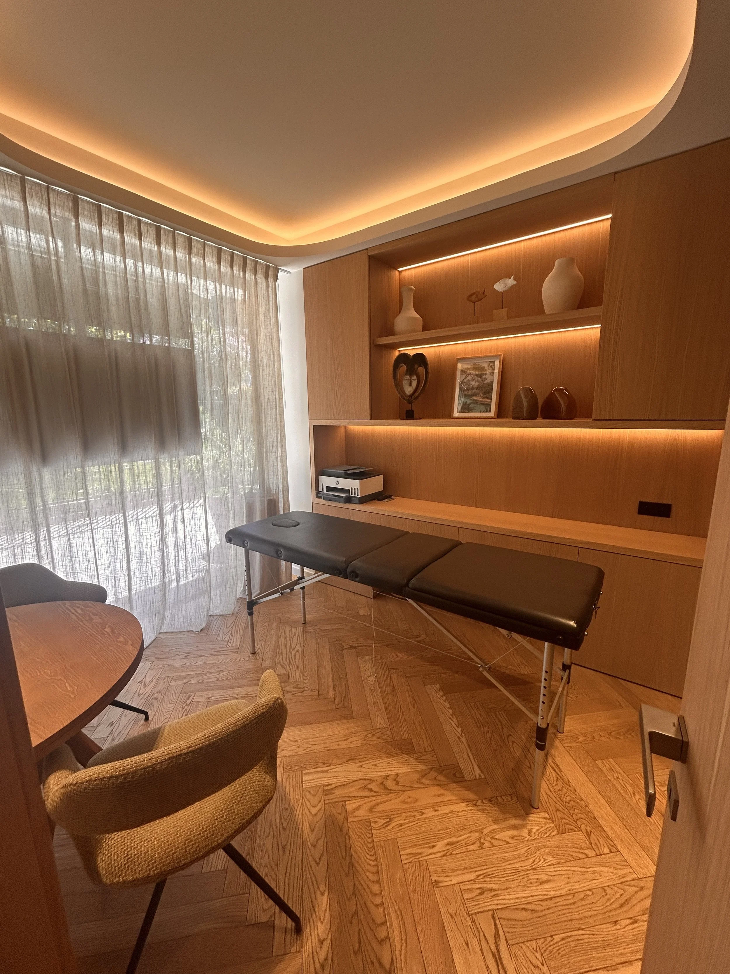 Une pièce meublée avec une table ronde, un fauteuil en osier, un lit de massage, des étagères en bois avec des décorations, et des rideaux transparents laissant entrer la lumière naturelle.