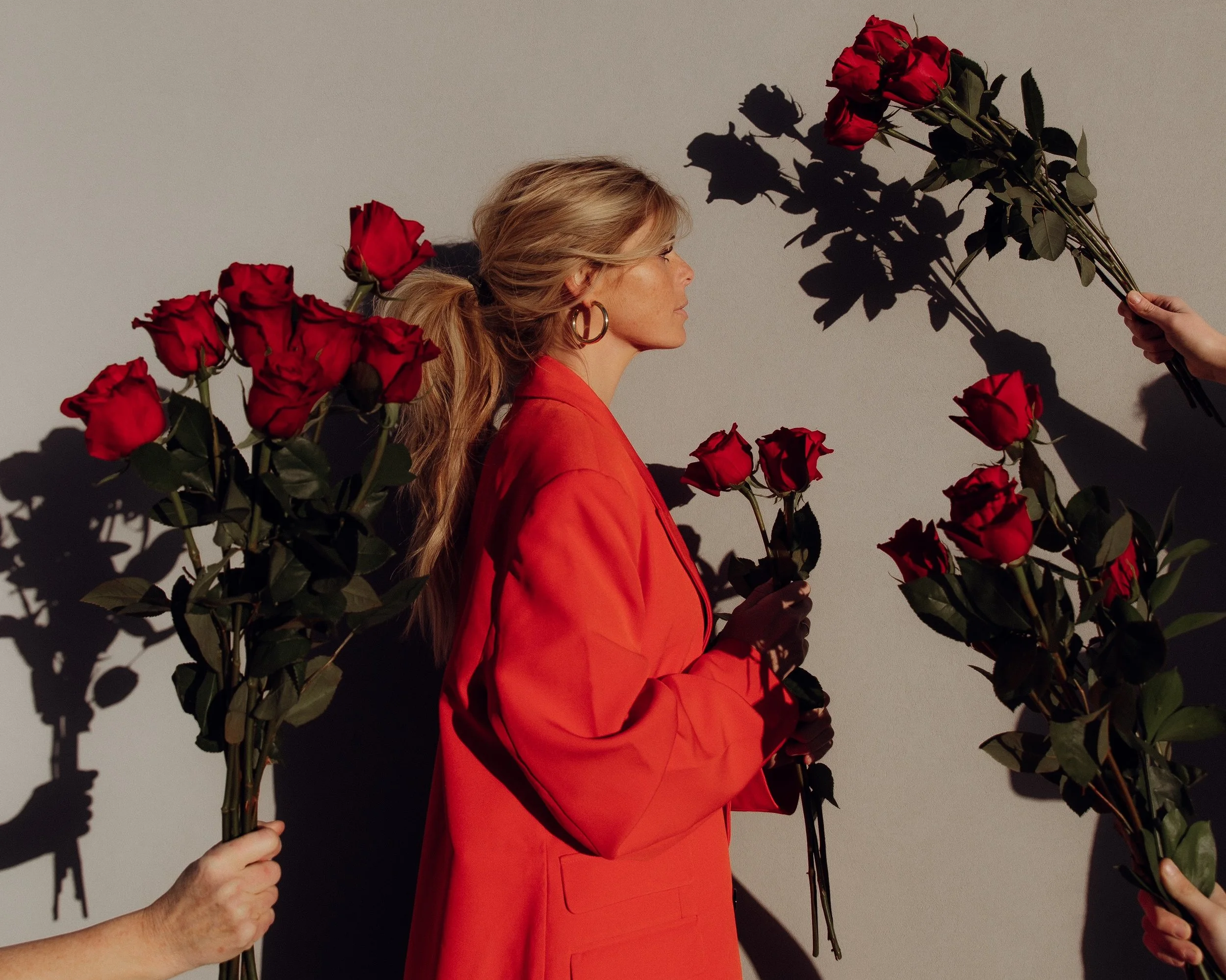 A woman with blonde hair, wearing a red blazer, stands sideways holding a single red rose. Several hands extend toward her, holding more red roses, creating a heart-shaped shadow pattern on the wall behind her.