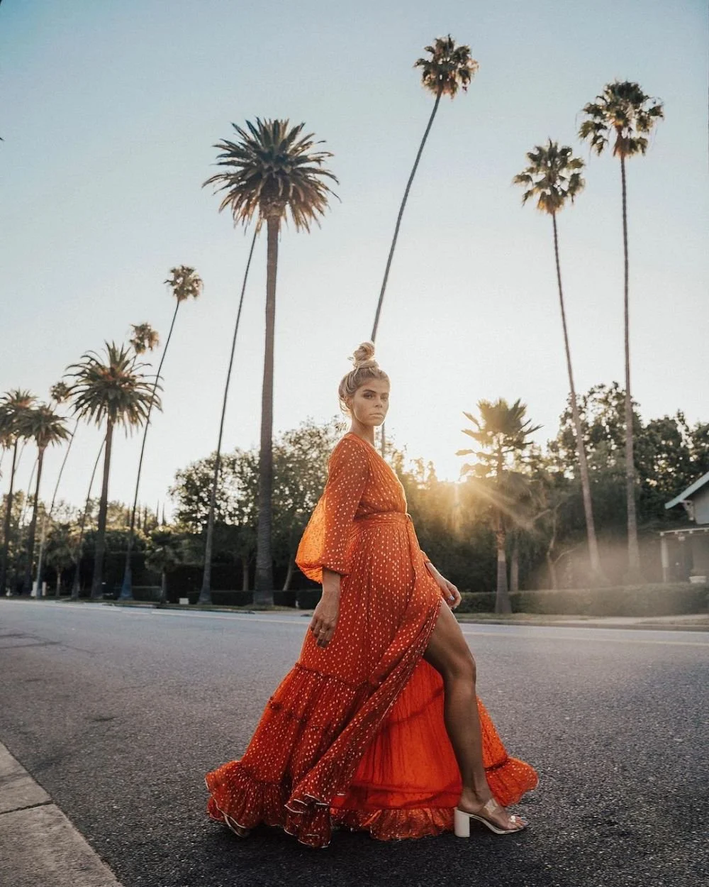 A woman in a long, flowing orange dress with a slit, standing on an empty street at sunset, with tall palm trees in the background.