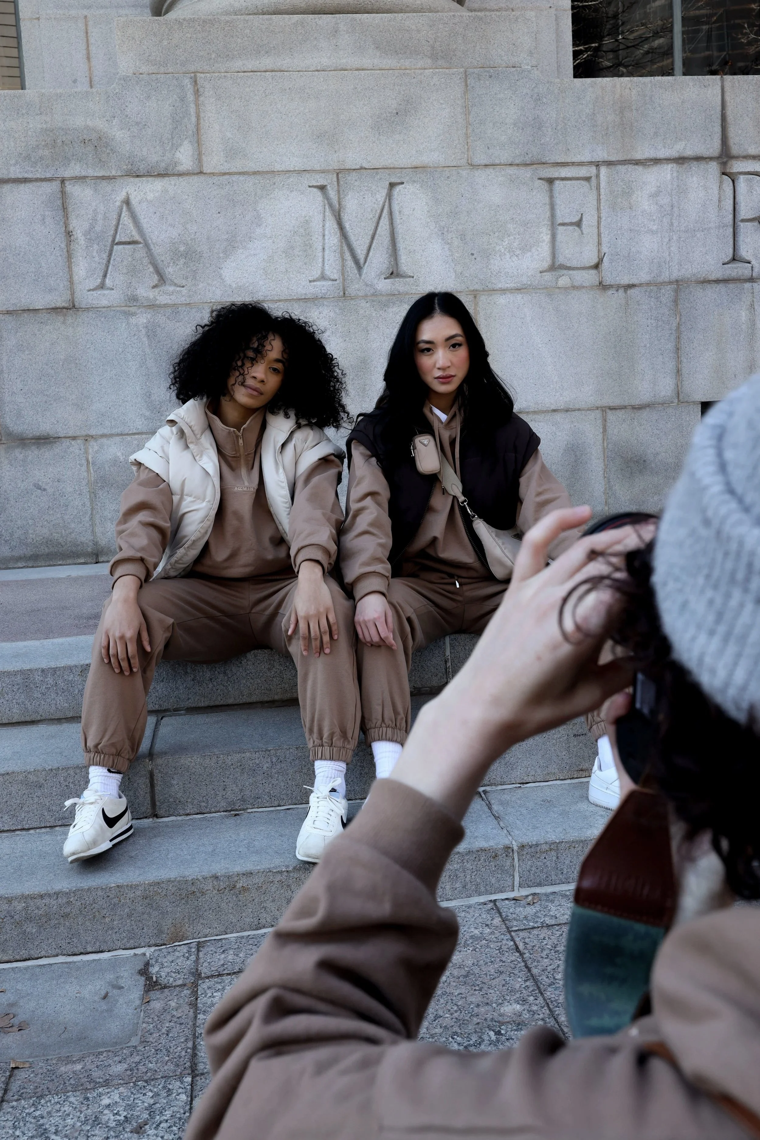 Two women sitting on stone steps posing for a photo, woman on left has curly hair and light-colored jacket, woman on right has straight hair and dark vest, both dressed in brown and white sneakers, with a photographer taking their picture in front of a stone wall with engraved letters.