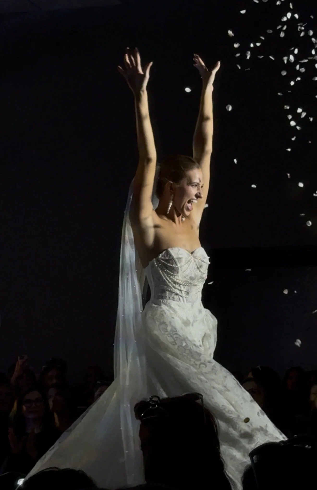 A woman in a wedding dress celebrating, with her arms raised and smiling, as confetti falls around her.
