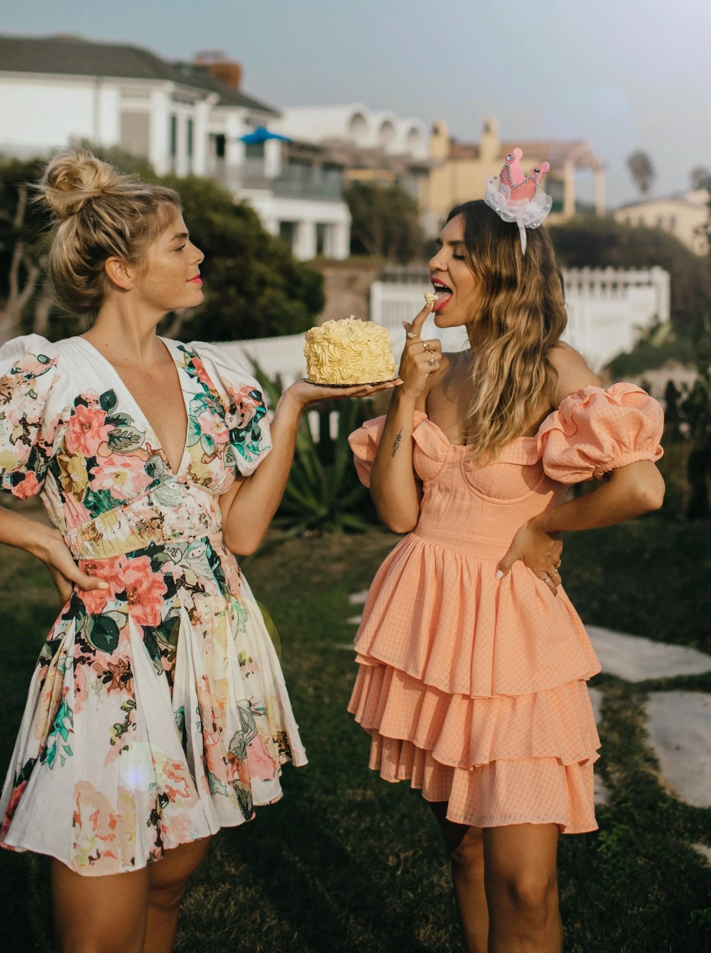 Two women in colorful dresses celebrating outdoors, one wearing a pink birthday crown, with a birthday cake in hand, in a suburban neighborhood.