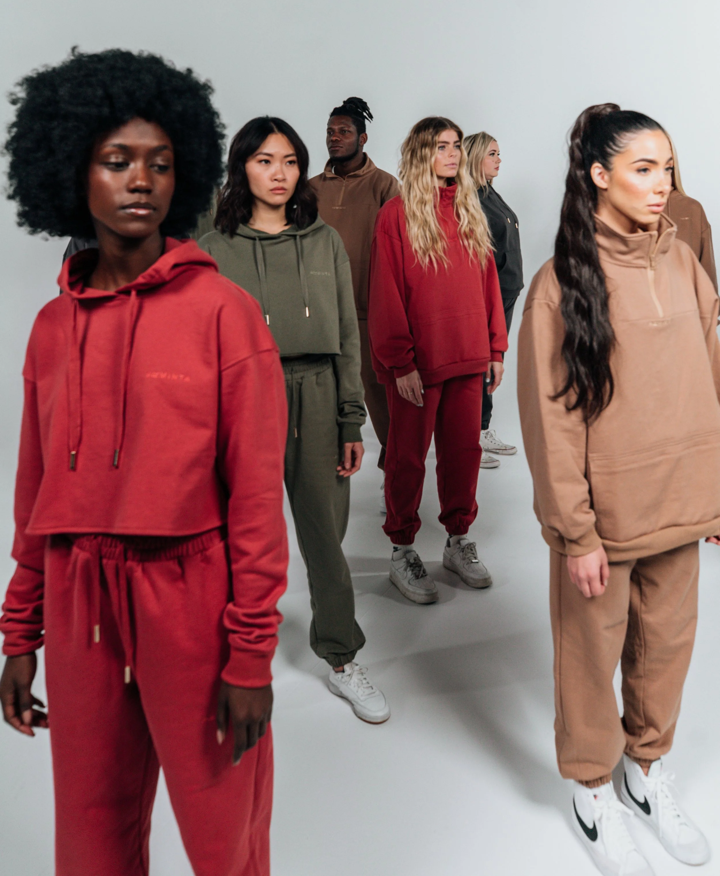 A group of seven diverse women standing in a white studio, dressed in colorful casual hoodies and sweatpants.