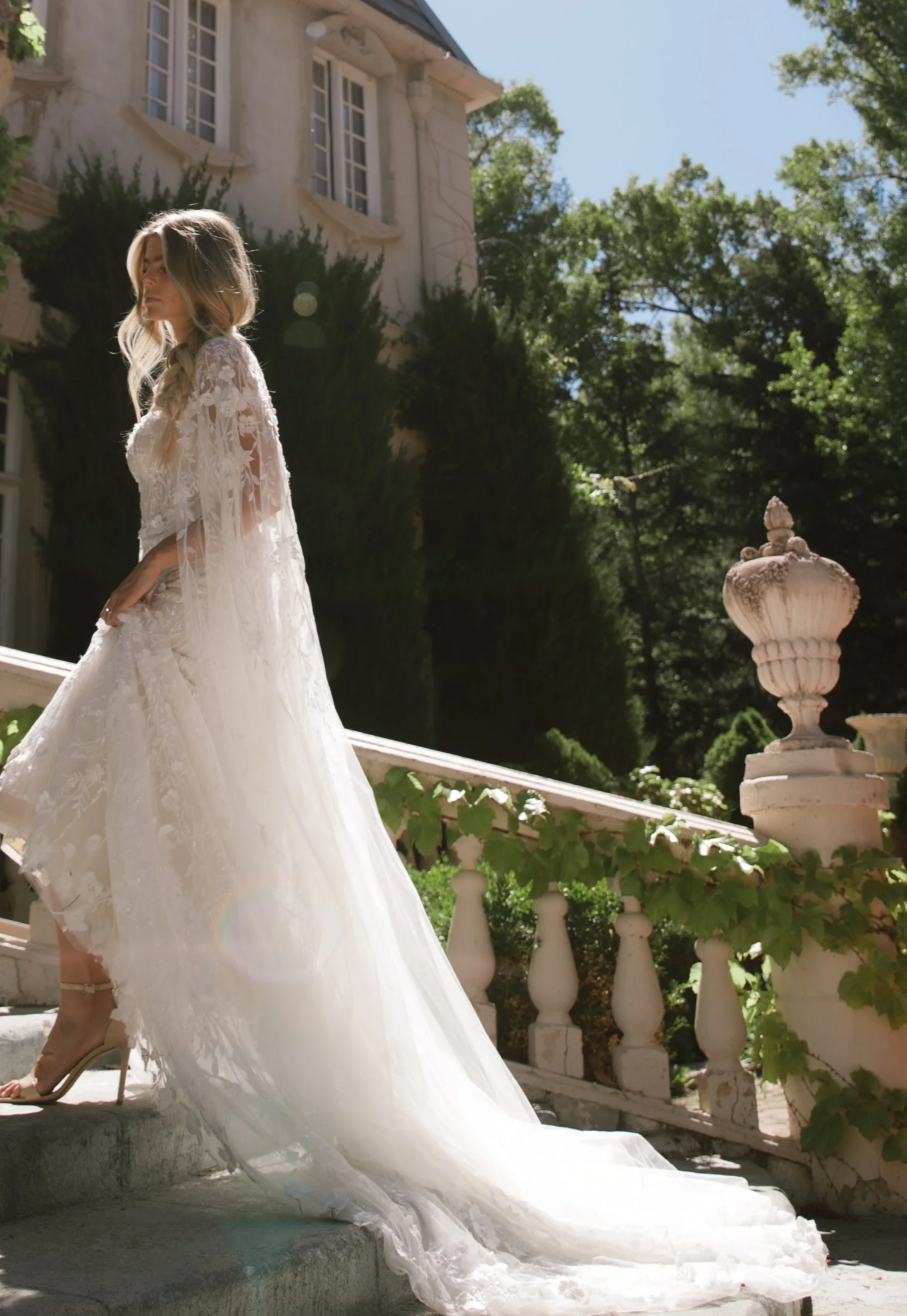 A woman in a white lace wedding dress and high heels standing on outdoor stone steps in front of a stately house with large windows, surrounded by lush green trees and foliage.