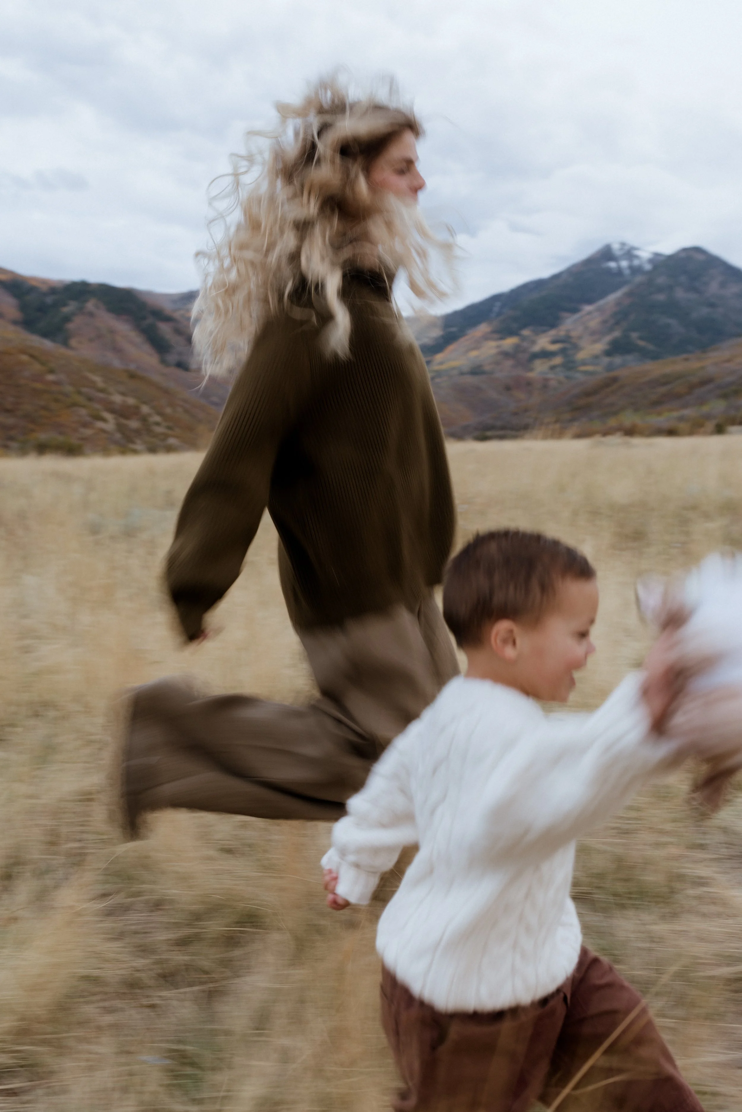 A woman with curly blonde hair and a young boy with short brown hair running through a grassy field with mountains in the background on a cloudy day.