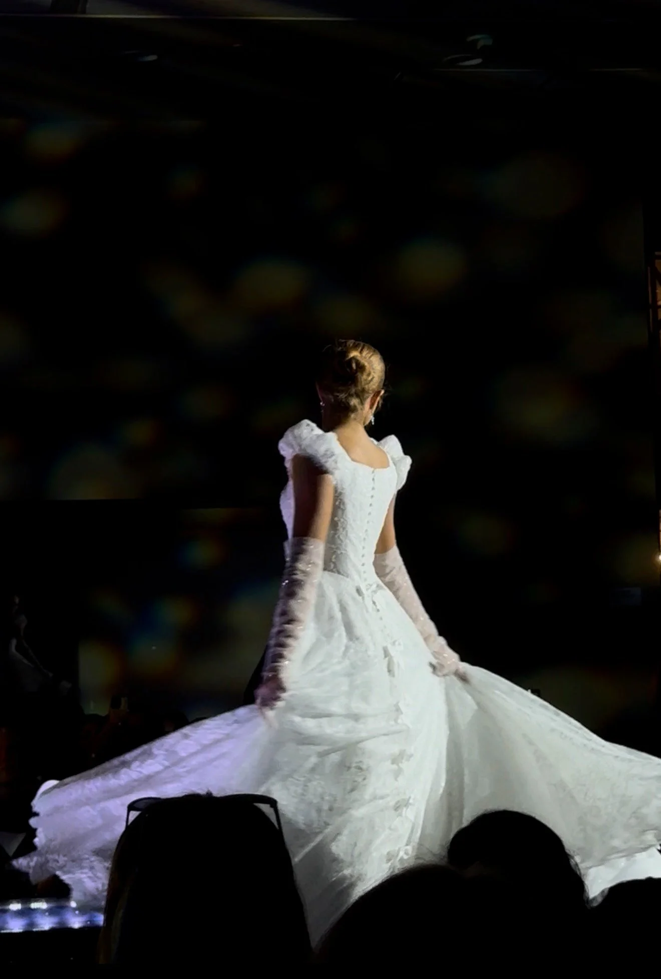 A woman in a white wedding gown and long gloves walking on stage with a dark background.