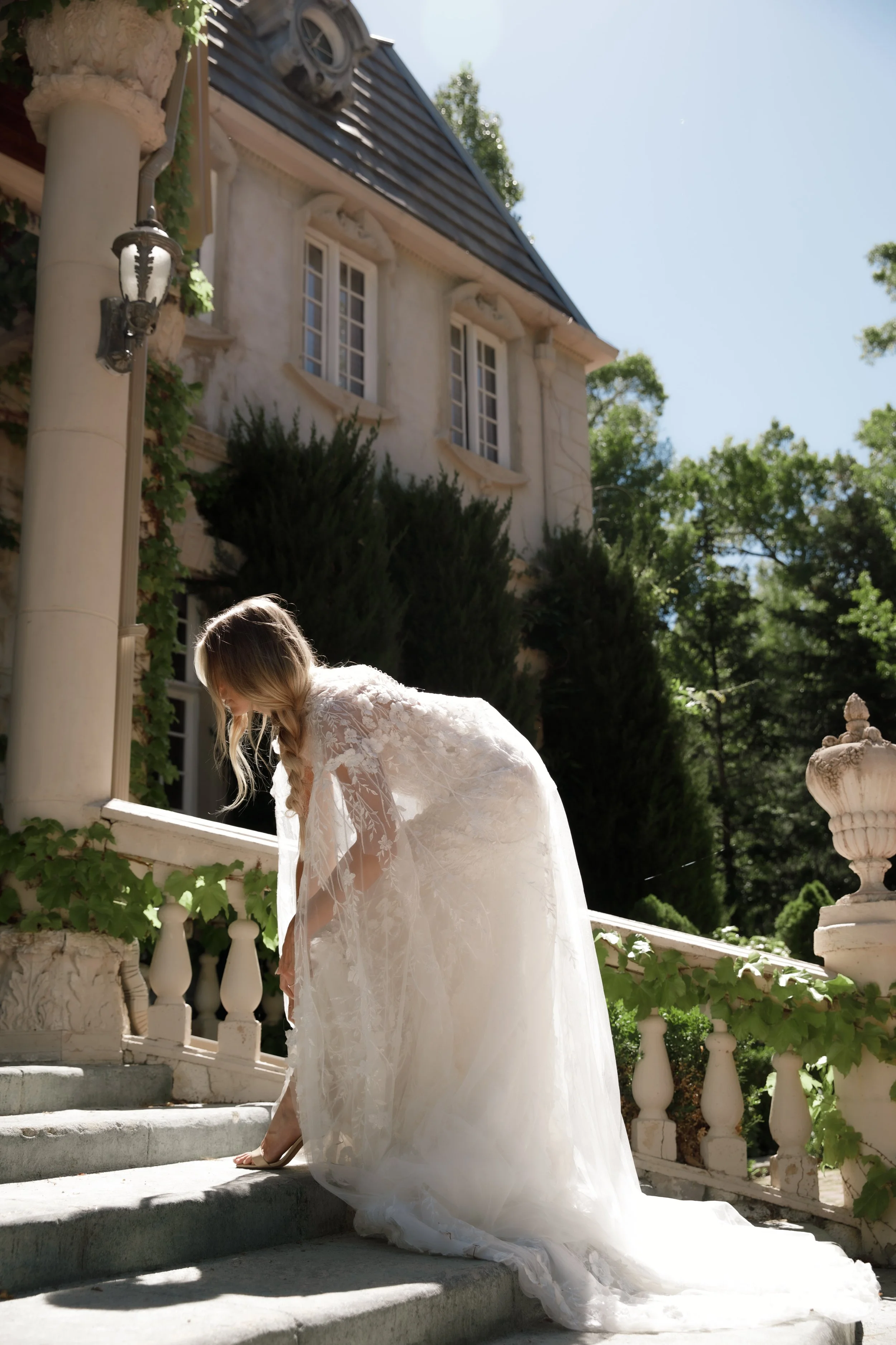 A woman in a wedding dress standing on steps outside a large, elegant house surrounded by trees.