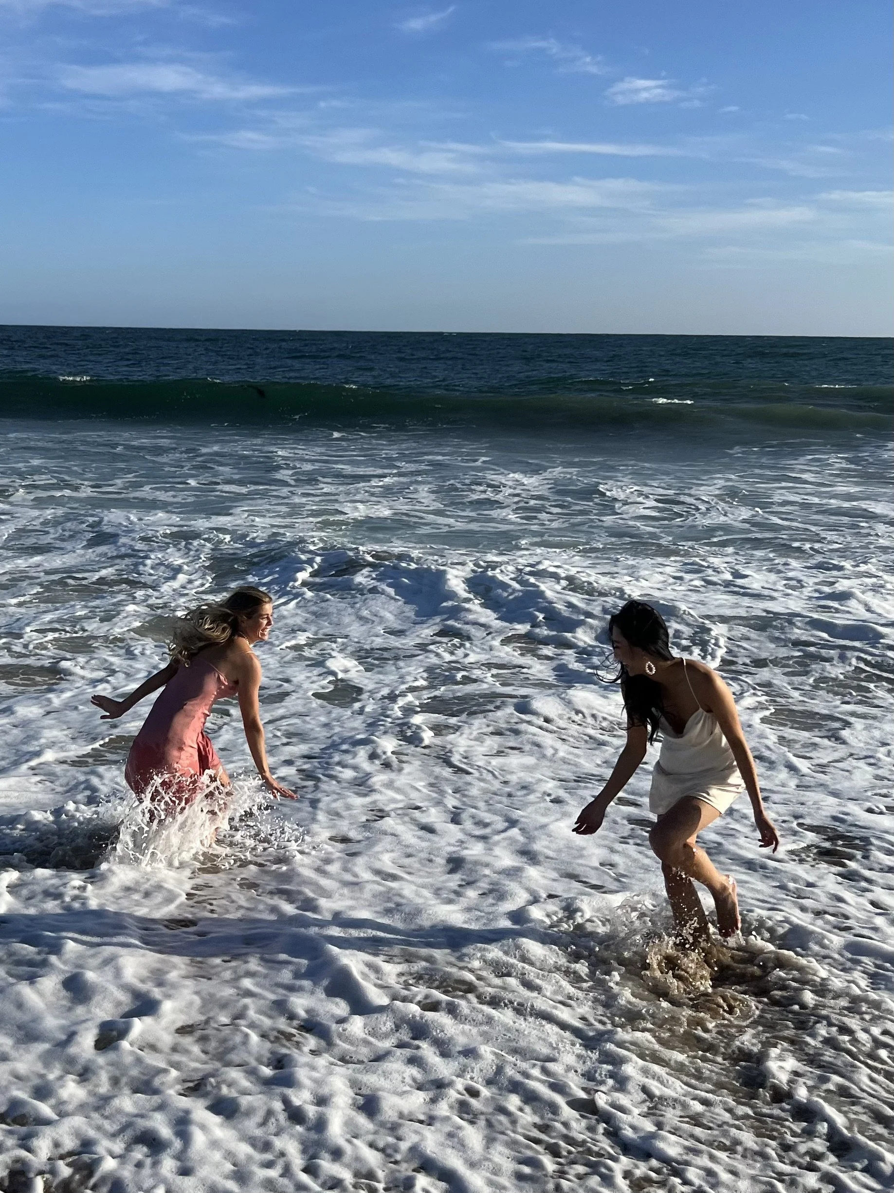 Two women playing in the surf at the beach, smiling and enjoying the water, with blue sky and ocean waves in the background.