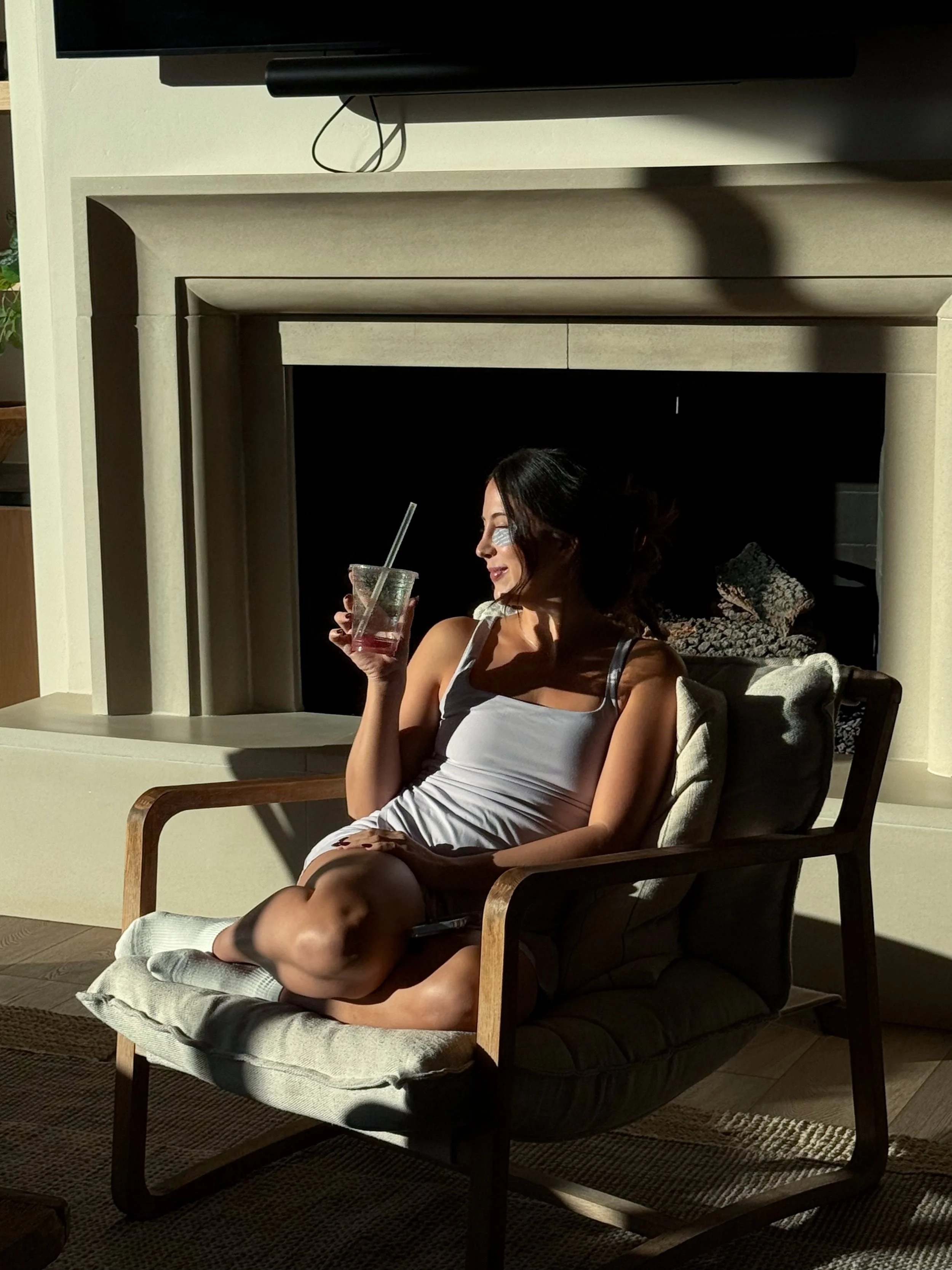 Woman sitting on a cushioned armchair holding a drink with a straw, illuminated by sunlight in a cozy living room with a fireplace and television.