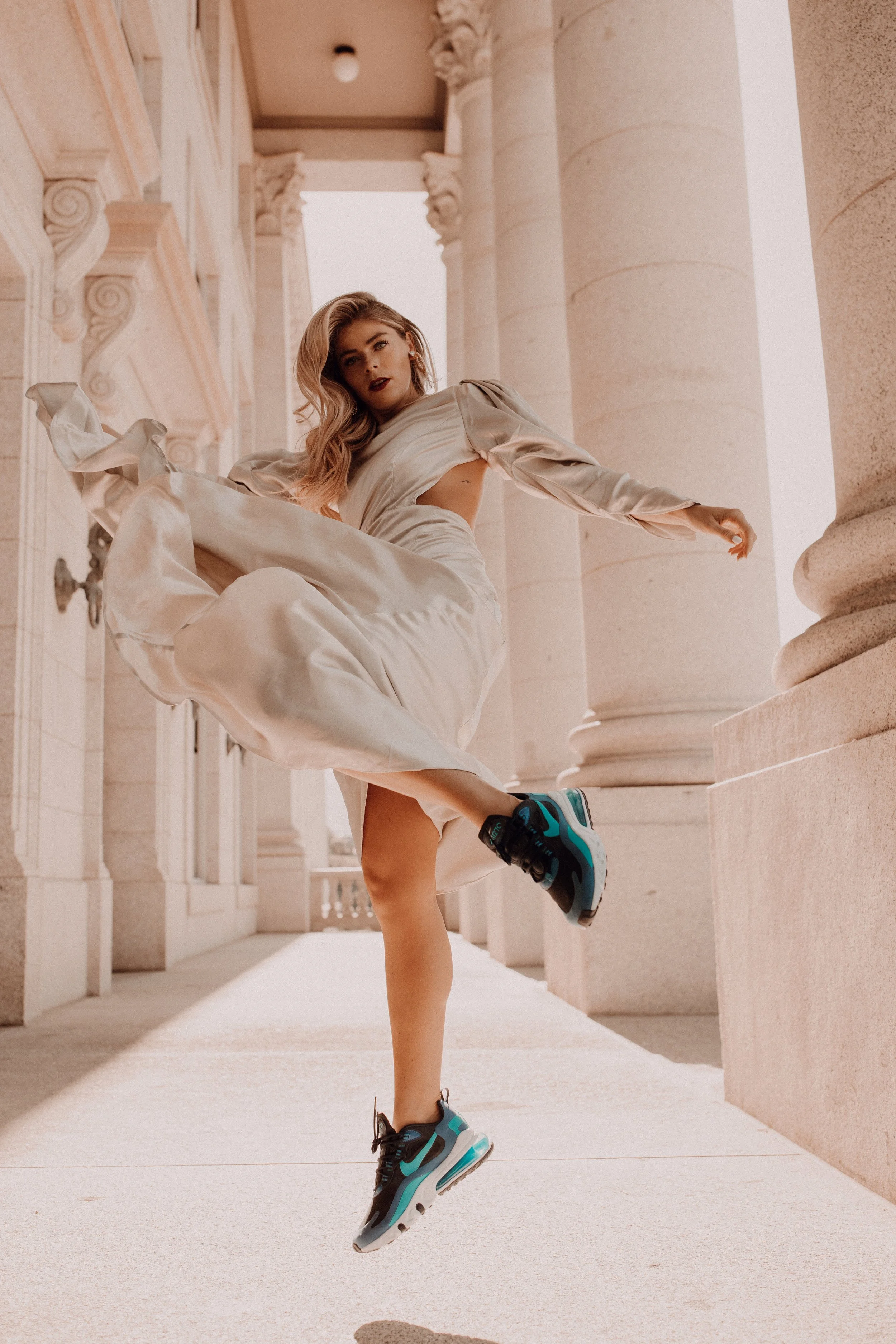 A woman jumping in the air in front of classical columns, wearing a long beige dress and black and turquoise sneakers.
