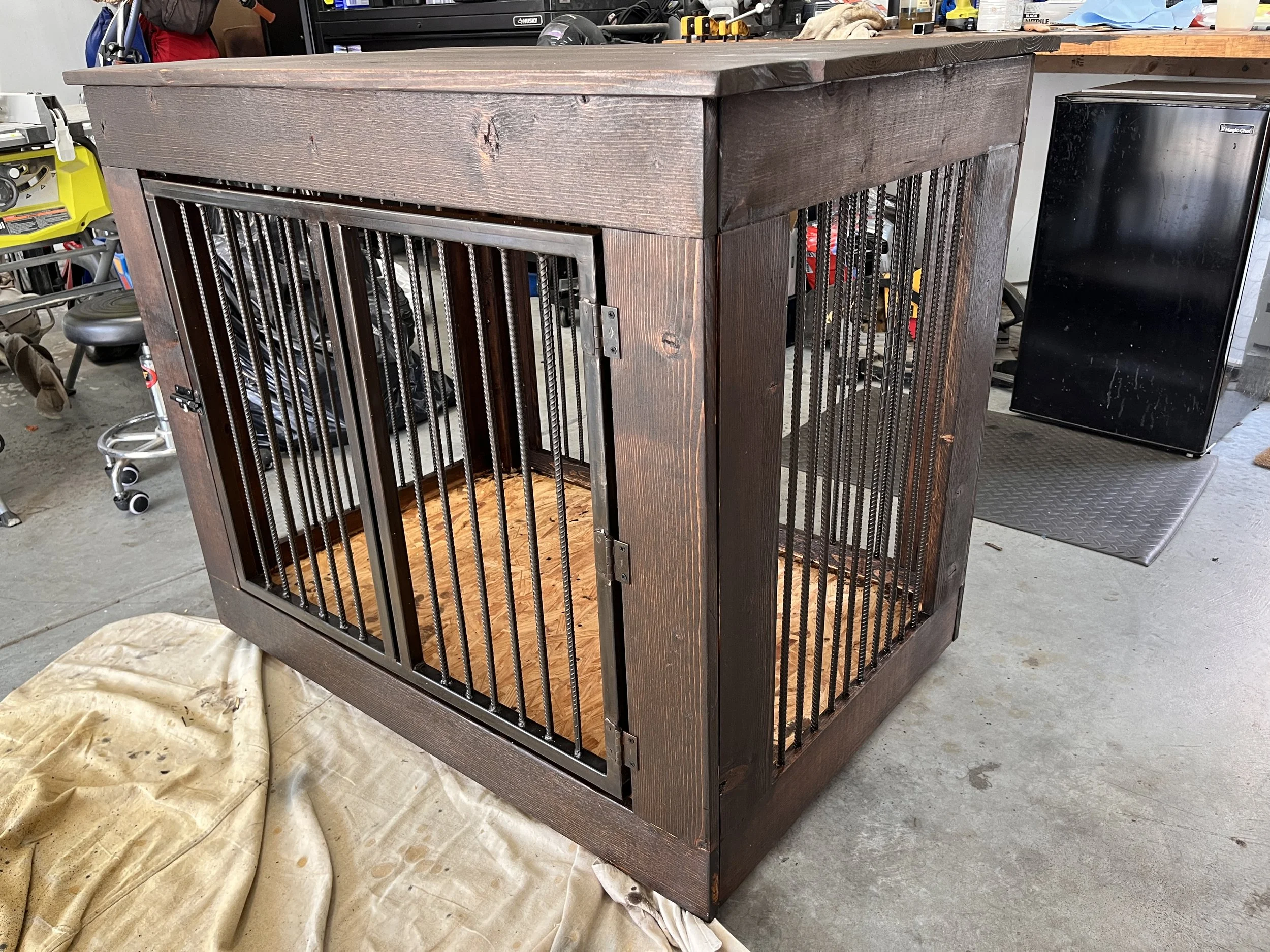 A wooden pet cage with metal bars on all sides, placed on a protective cloth on the floor in a workshop or garage setting. The background includes various tools and equipment.