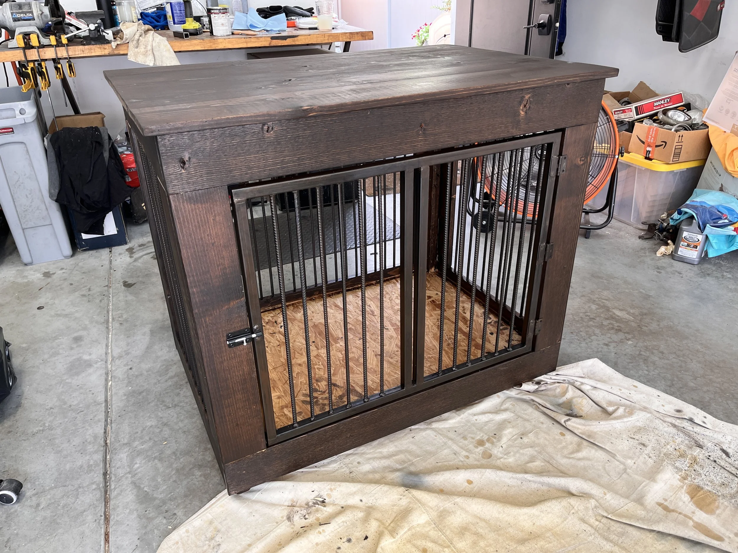 A wooden dog crate with black metal wires for the door, placed in a garage or workshop. The crate has a dark stain finish and a wooden base.