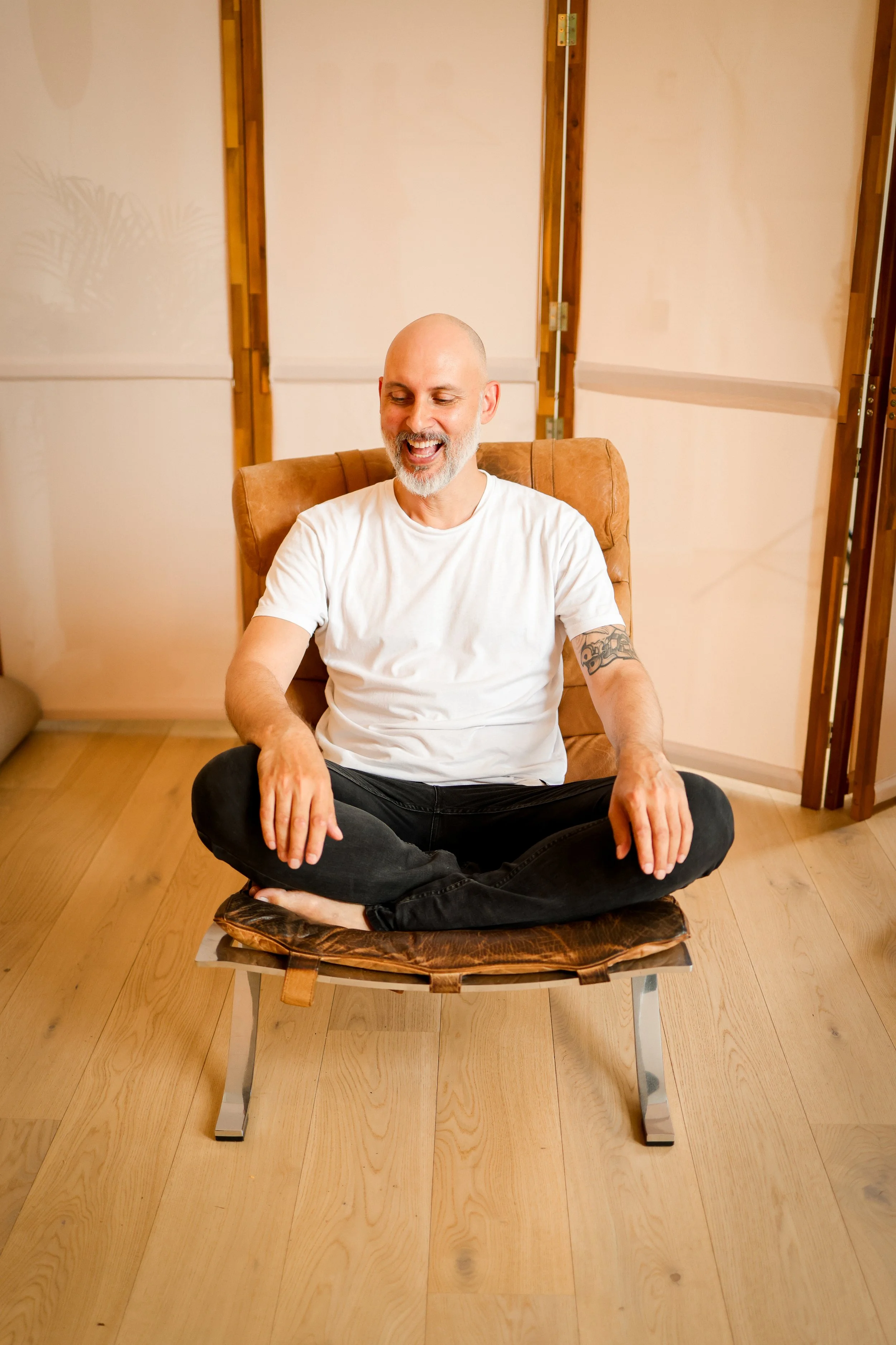 A man with a shaved head and gray beard, sitting cross-legged on a leather cushion on a wooden chair. He is wearing a white T-shirt and has a tattoo on his left arm. He appears to be meditating or relaxing with a smile on his face. The background features a beige wall with wooden framing and a wooden floor.