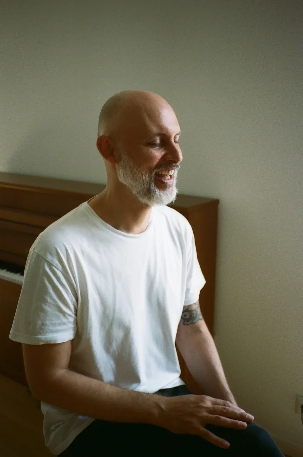 A middle-aged man with a bald head, white beard, wearing a plain white T-shirt, sitting with a piano in the background, smiling with eyes closed.