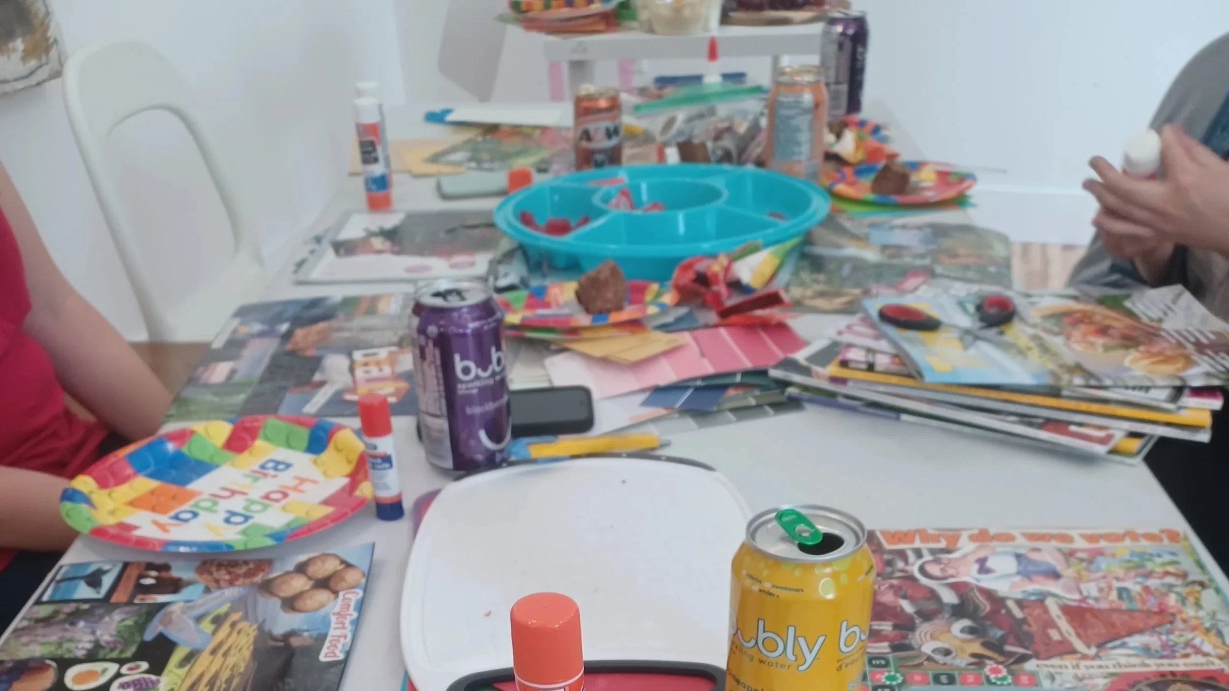 A cluttered table with collage materials, magazines, scissors, glue sticks, and a small blue plastic container, with people partially visible around it working on collages.
