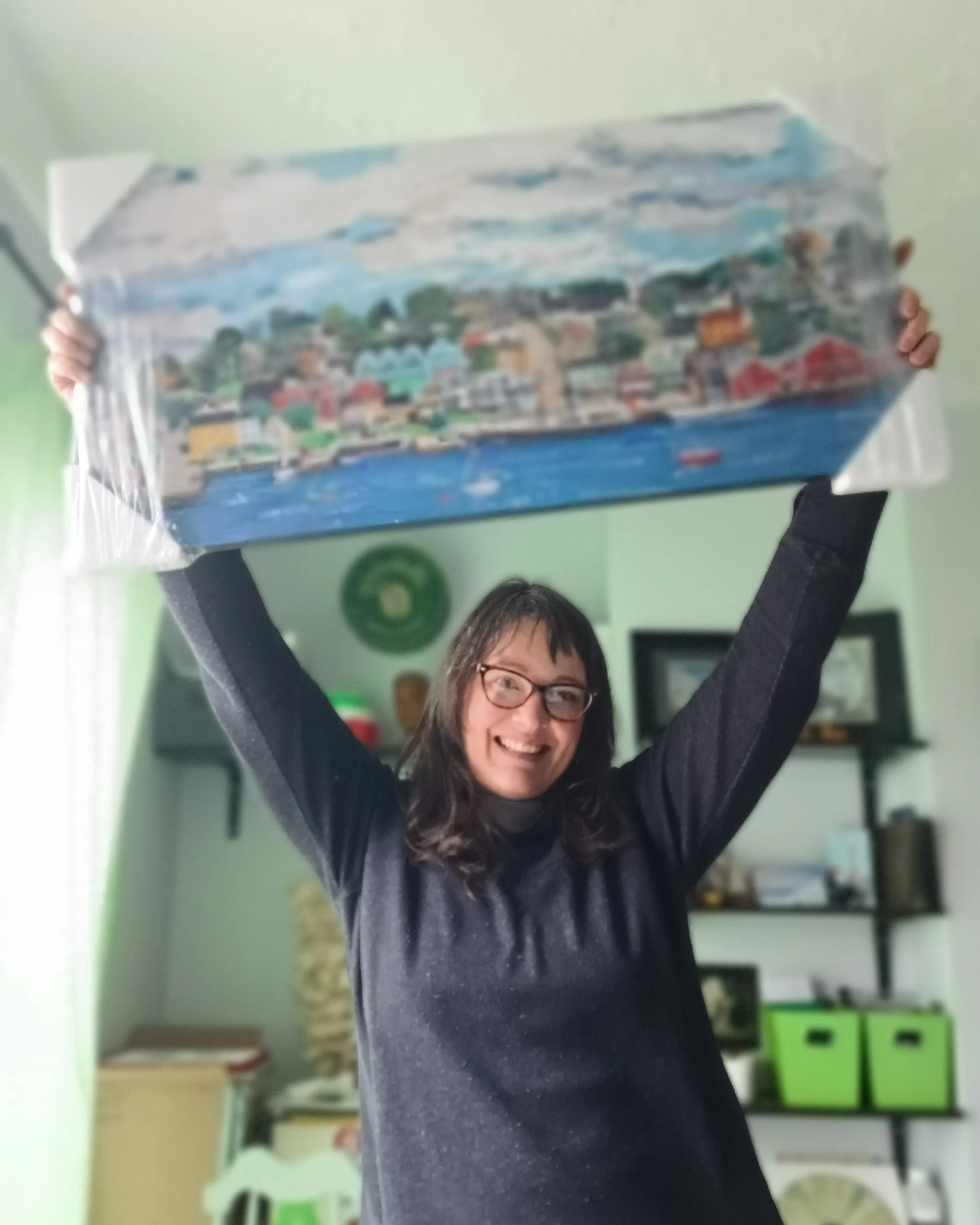 A woman with glasses smiling and holding a large, colorful model of a town or cityscape above her head in a room with green walls and shelves with various objects.