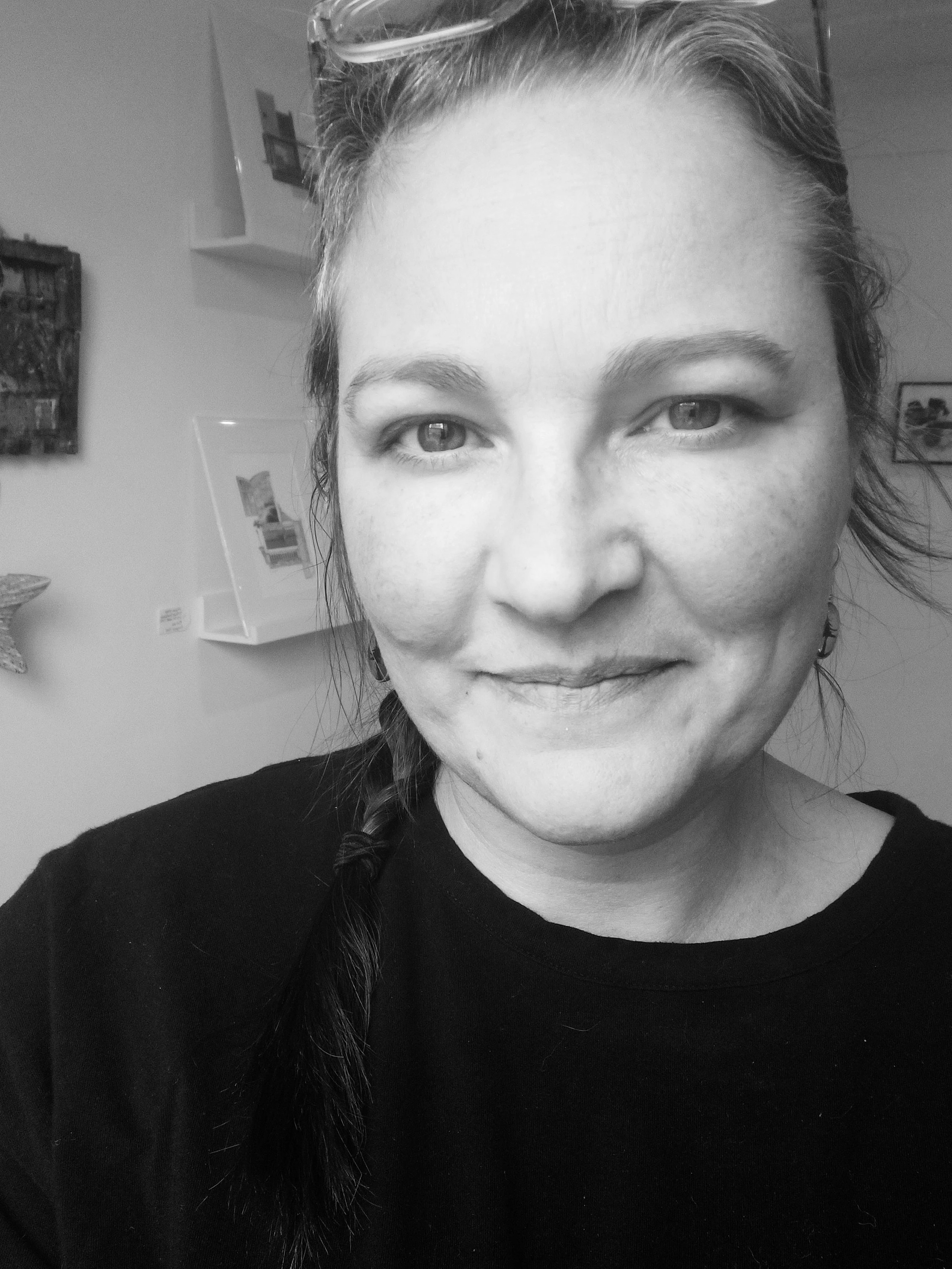 A woman with glasses on her head, smiling, with her hair in a braid, wearing a black shirt, taking a selfie in front of a wall with framed pictures and artwork.