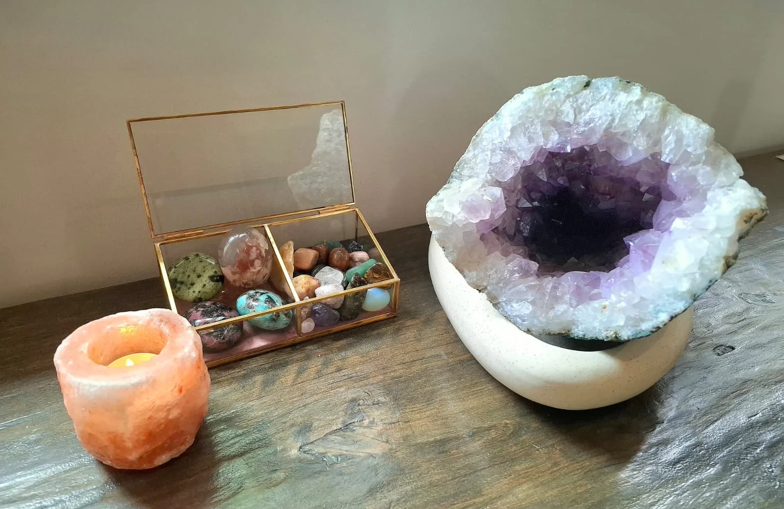 Een houten tafel met een glaskist vol verschillende gekleurde edelstenen, een grote geode met paarse kristallen erin, en een kleine theelicht in een roestkleurige houder.