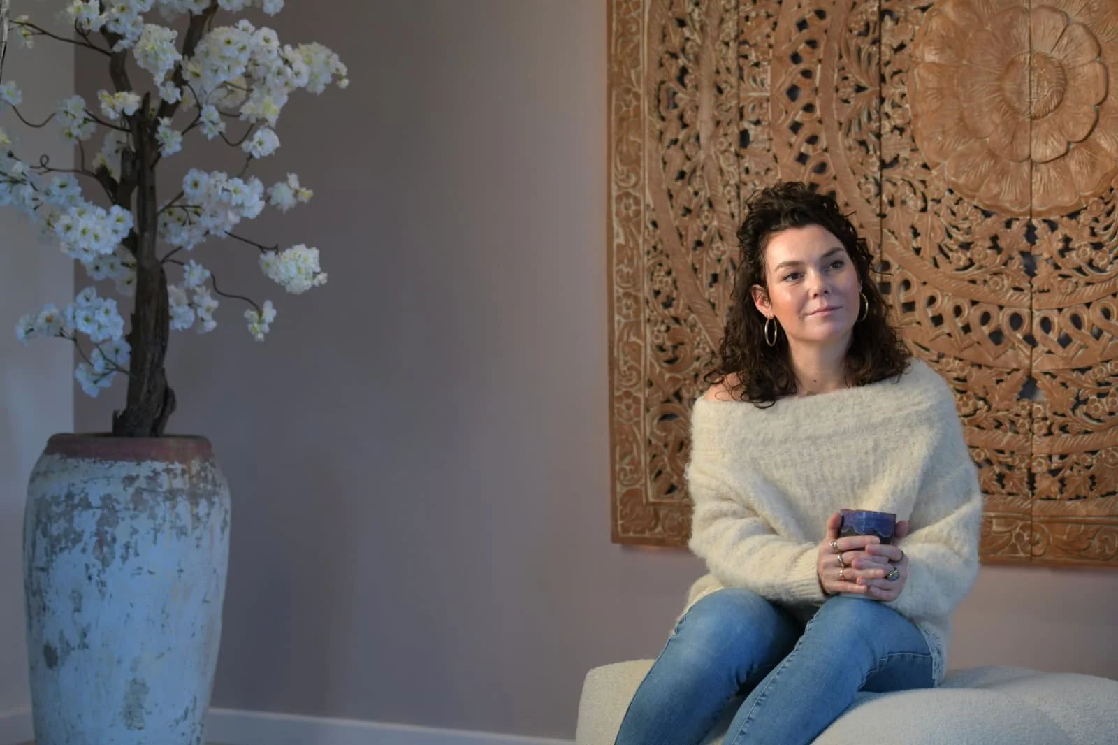 Vrouw zit op een comfortabele bank, houdt een mok in beide handen, draagt een zachte beige trui en blauwe jeans, met een houten wanddecoratie achter zich en een grote witte bloempot met kunstbloemen links.