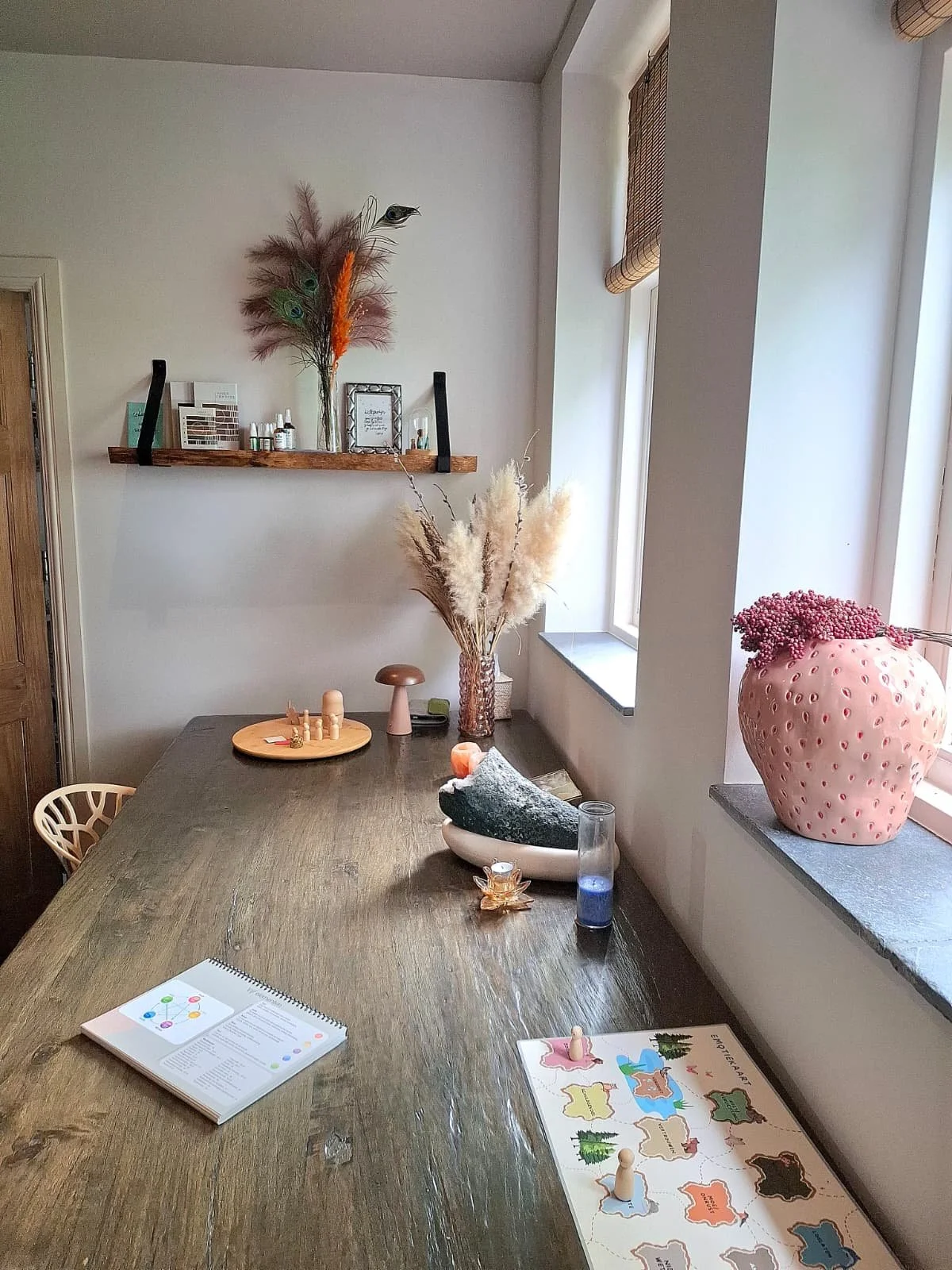 Een houten tafel met een bordspel en een boek, naast een vaas met pampasgras en decoratieve objecten, bij een raam met een roze vaas met bloemen.