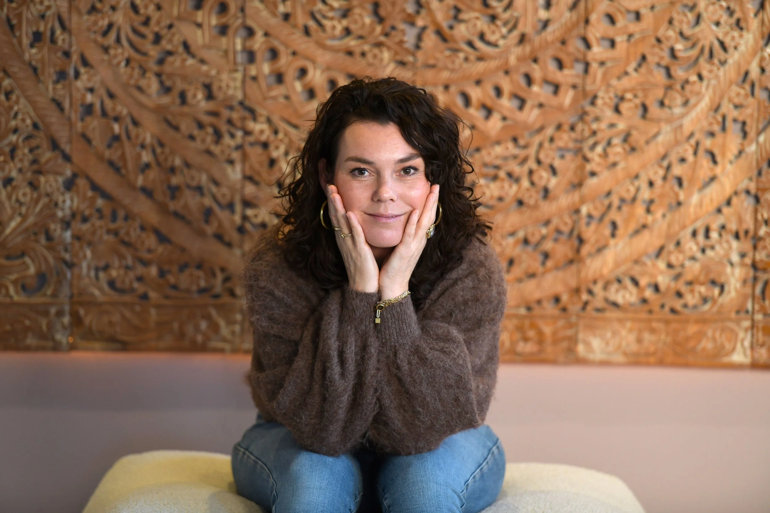 Een vrouw met krullend donker haar, goudkleurige oorbellen en sieraden, zittend op een gele poef voor een gedetailleerd houtgesneden kunstwerk op de achtergrond.