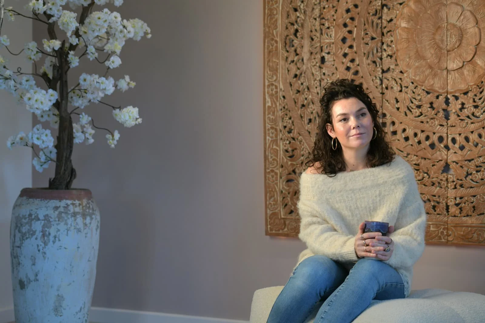 Vrouw zit op een bankje, houdt een mok vast en kijkt naar voren. Op de achtergrond een gedetailleerd houten wanddecor en links een grote witte vaas met een kunstboom met witte bloemen.