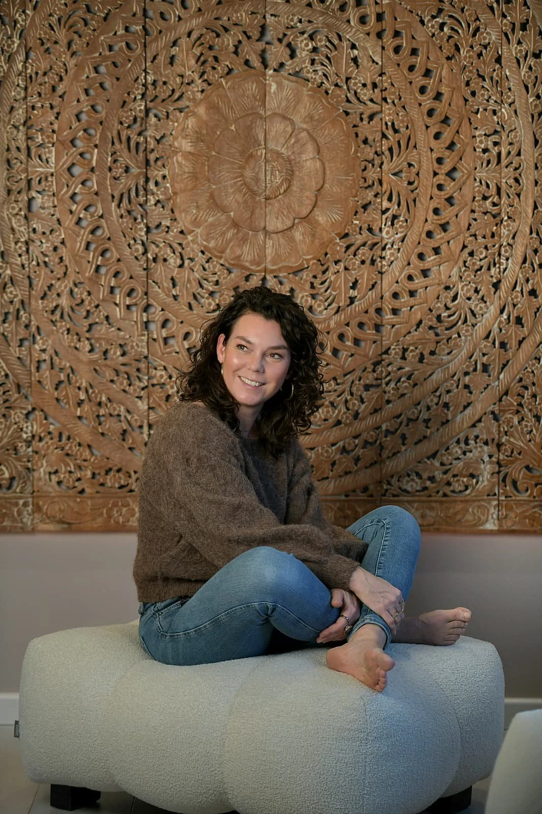 Een vrouw zit op een bordeauxkleurige bank met een grote decoratieve houten muur achter haar, haar benen gekruist, glimlachend, met vloeiende donkerbruine haar.