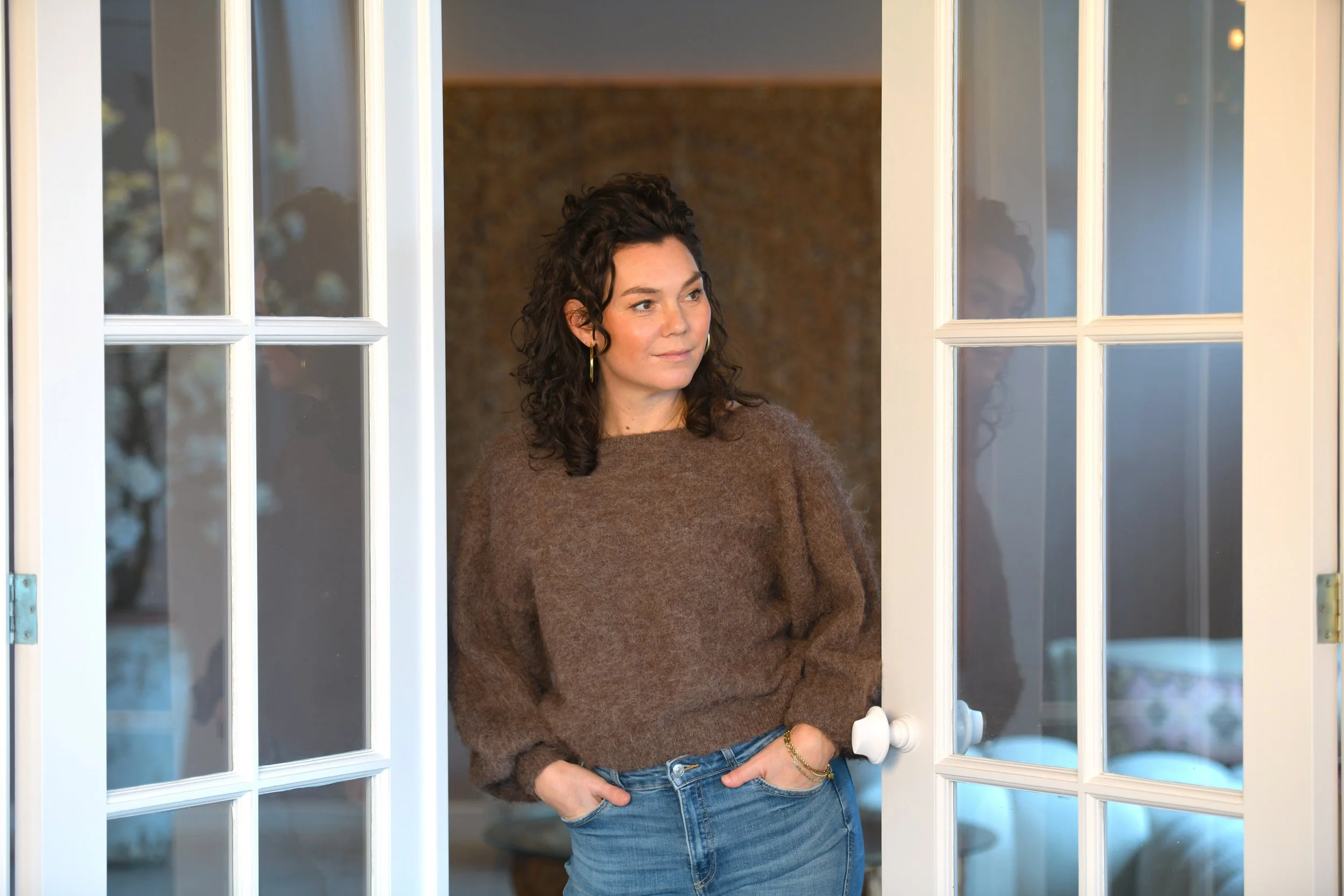 Vrouw staat voor een openslaande deur, met haar hand in haar zak, en kijkt naar links.