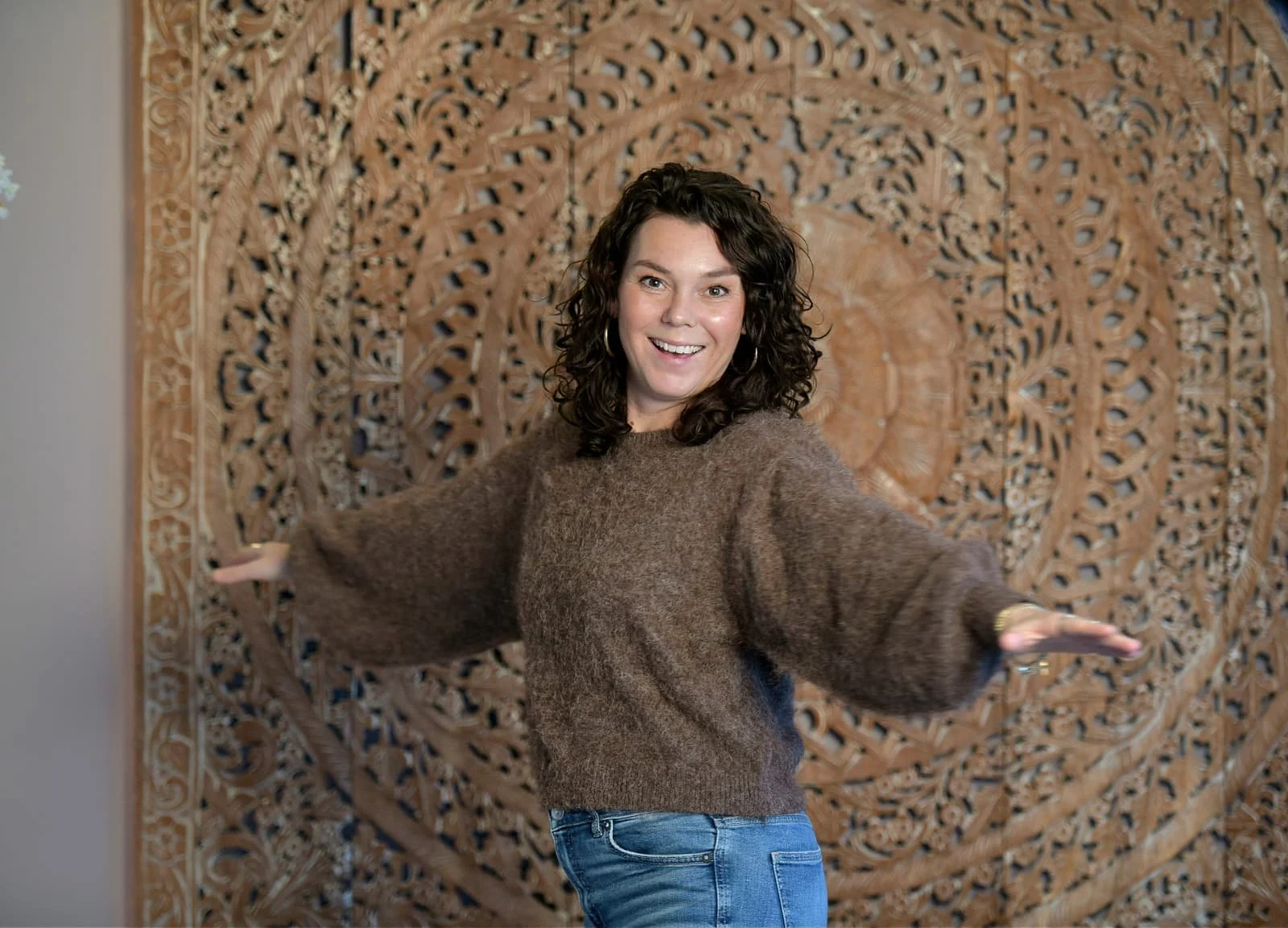 Vrouw met gekrulde haar in een bruine trui poseert voor een stenen muur met reliëf.
