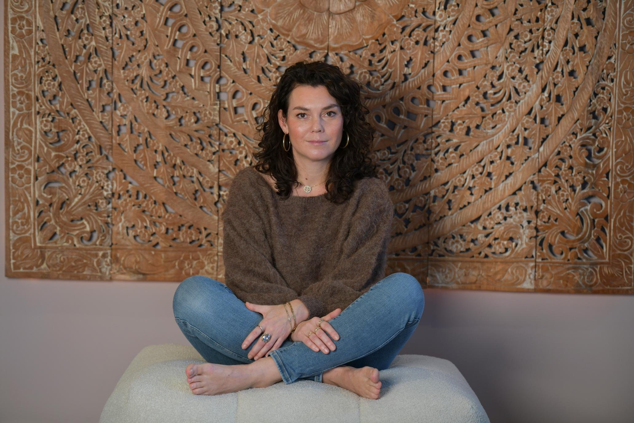 Vrouw zittend in lotushouding voor een intricaat houten kunstwerk op de achtergrond.