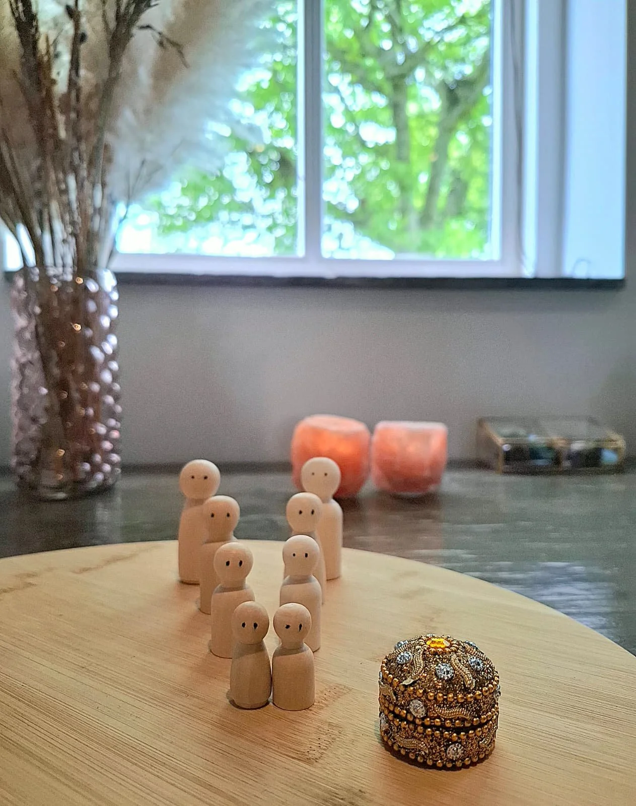 Een houten tafel met houten poppetjes, een sieradendoosje, kaarsen, een glazen pot met gedroogde bloemen, en een venster met groene bladeren buiten.