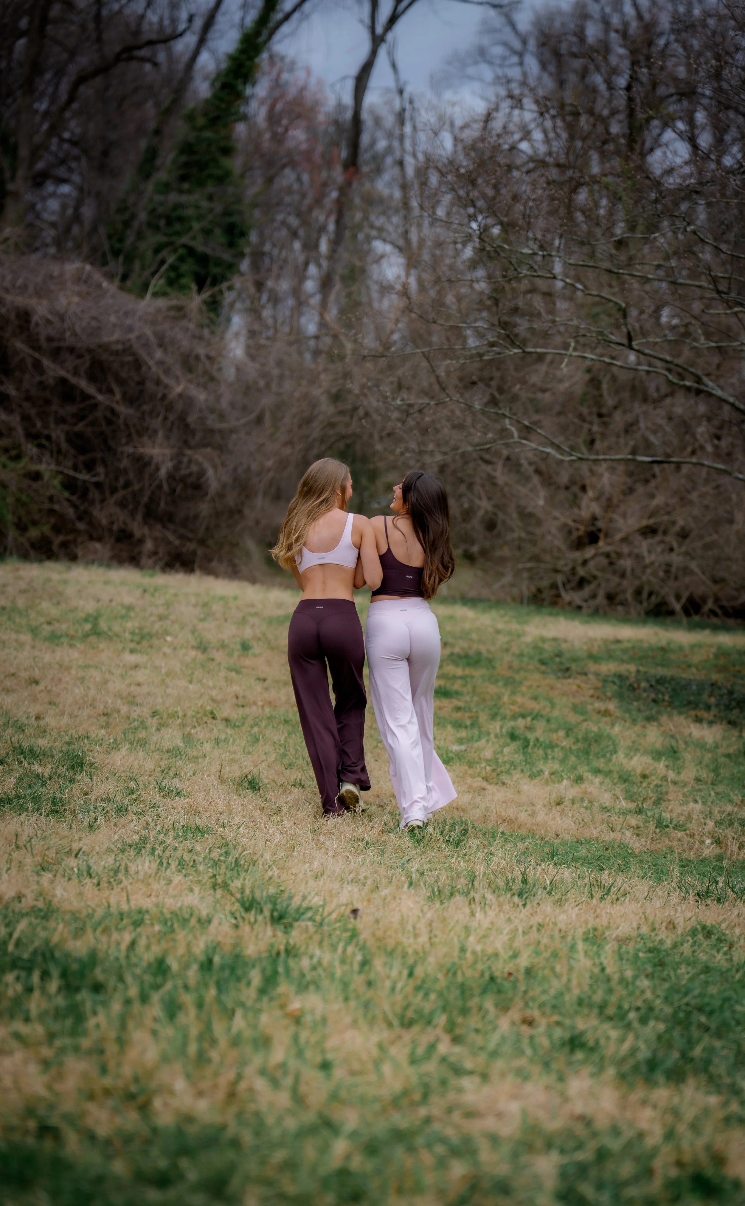 Two women walking together in a grassy park during autumn, surrounded by leafless trees.