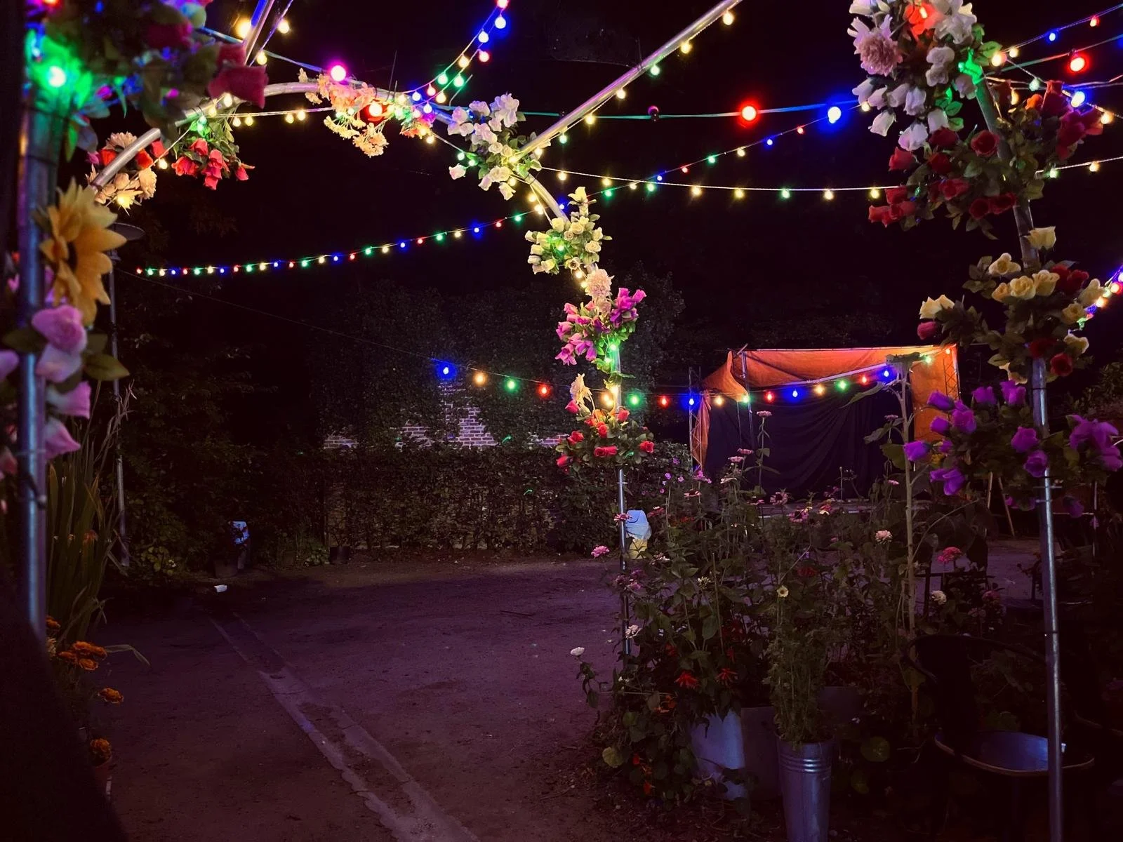 Nighttime scene of an outdoor event decorated with multicolored string lights and artificial flowers on metal frames, with a small stage and potted plants along the pathway.