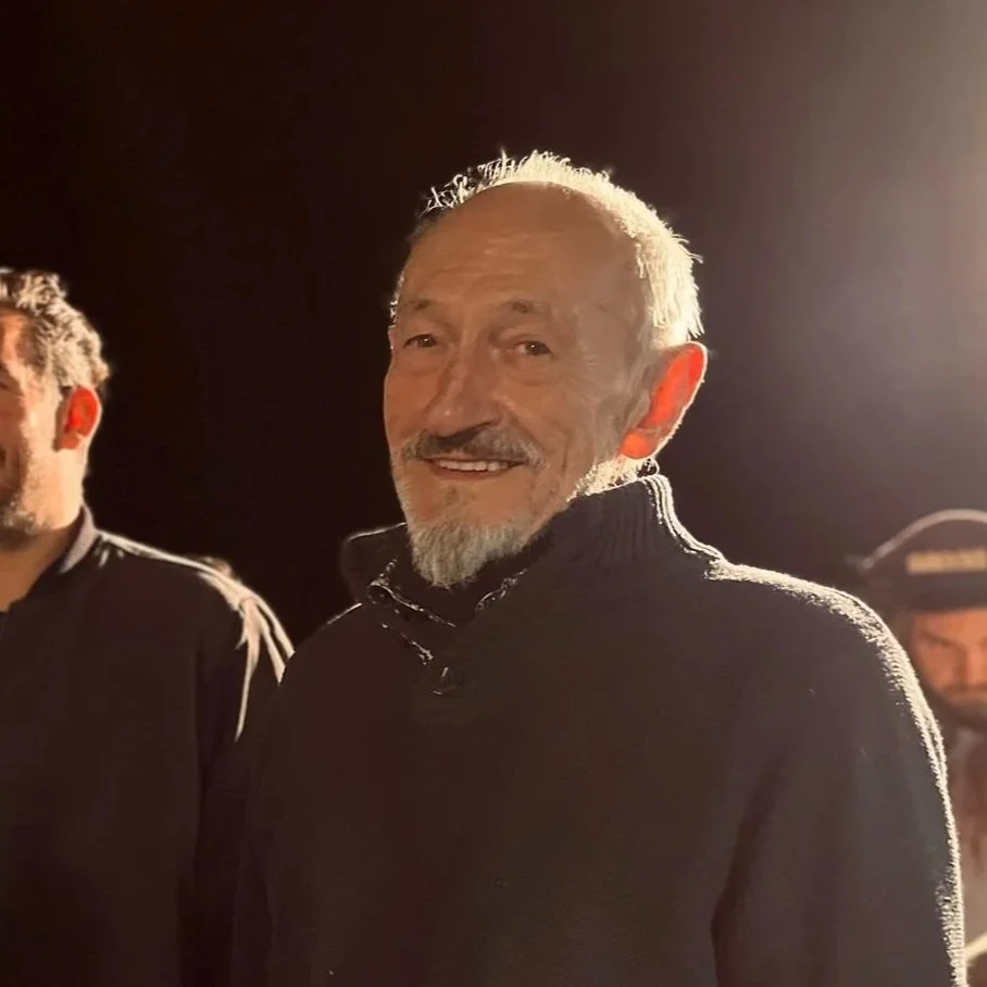An elderly man with white hair and a beard smiling, wearing a dark sweater, standing in a dimly lit setting with other people partially visible in the background.