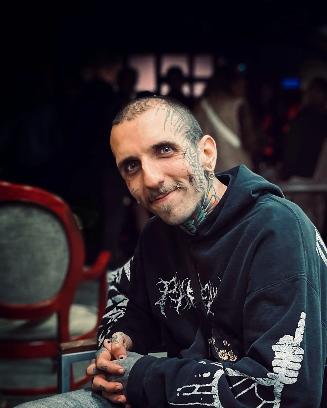A man with tattoos on his face and head, wearing a black hoodie, sitting at a table with a smile.