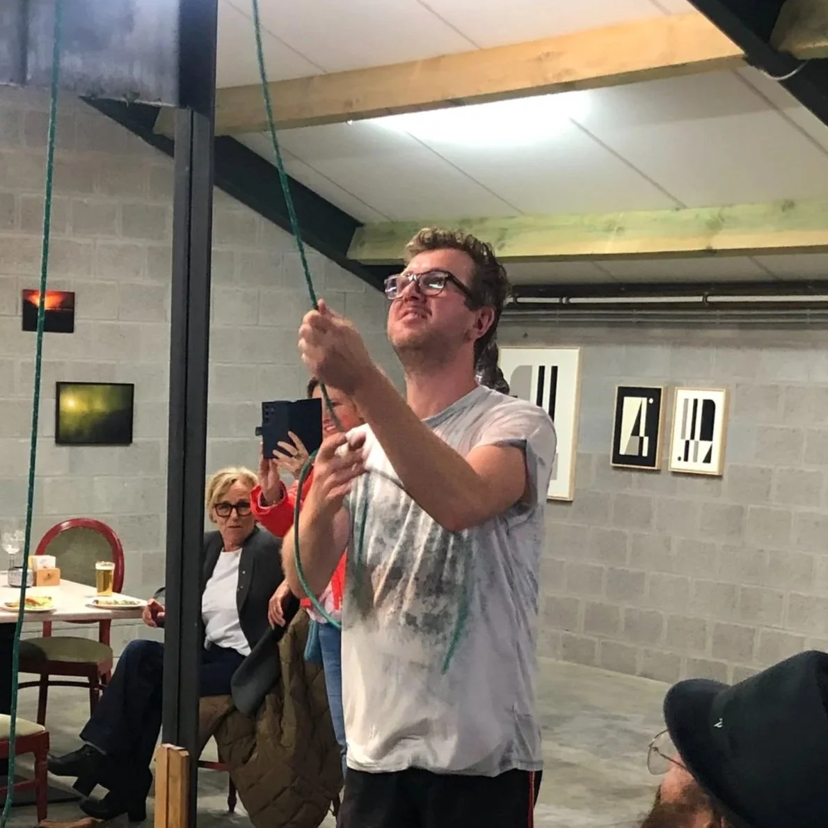A young man with glasses and curly hair, wearing a tie-dye t-shirt, is holding a rope or cord, looking at it intently, while a woman with glasses and short blonde hair, wearing a black jacket, is seated at a table in the background, with plates of food and drinks, and other people are present in an art gallery or exhibition space with framed artwork on the wall.