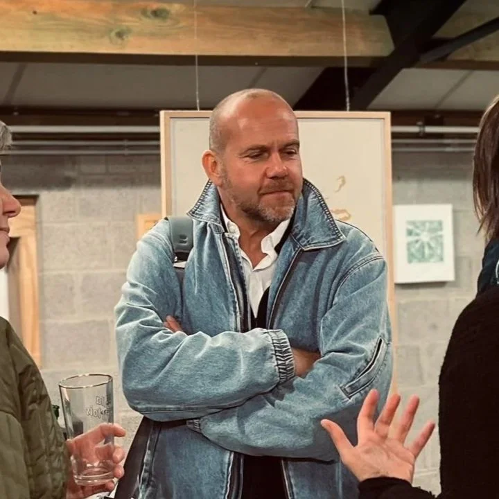 Three people engaged in conversation, with a man in a light blue denim jacket at the center, crossing his arms and listening, inside an art gallery or similar space.