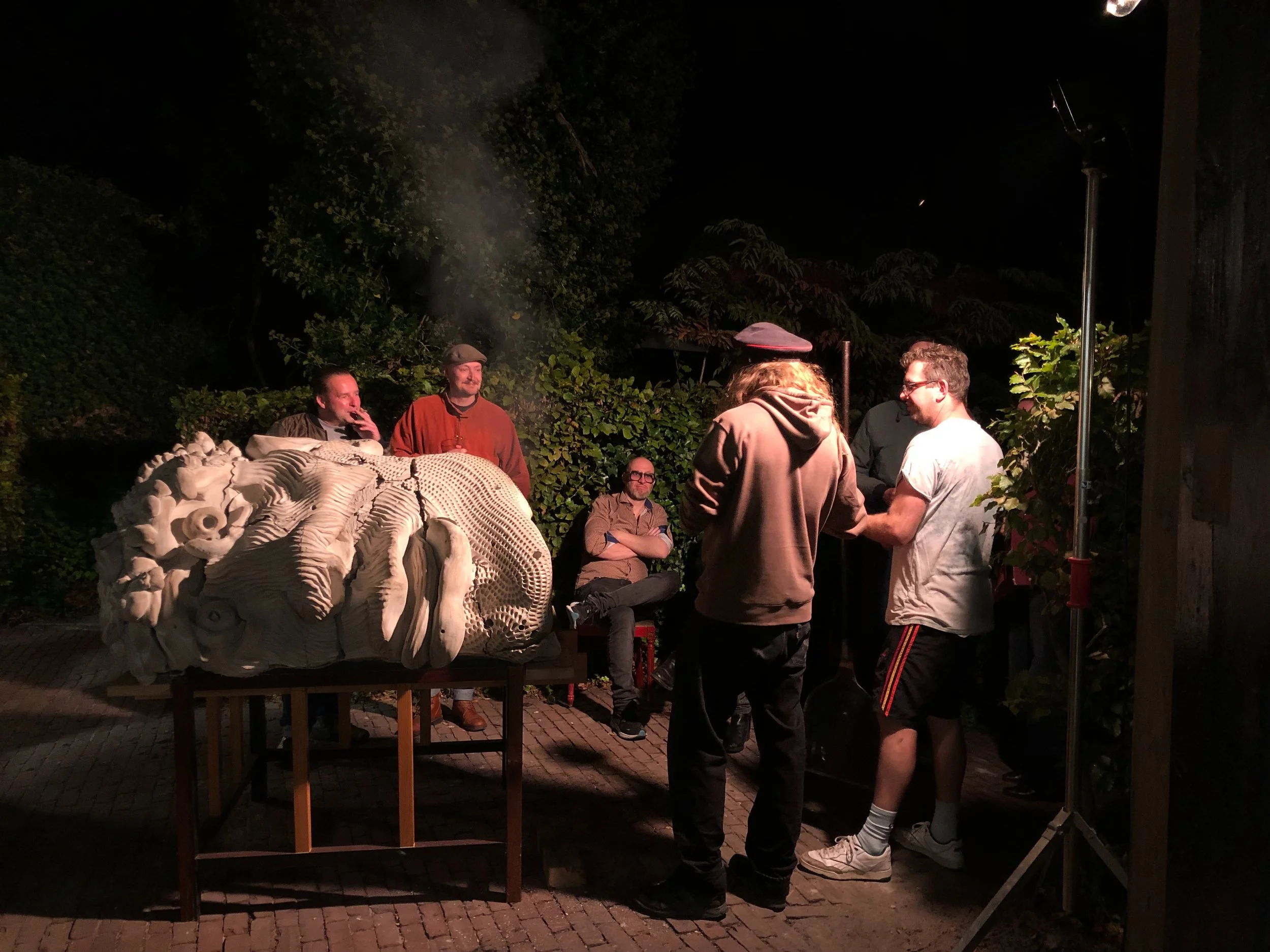 Group of people standing and sitting in an outdoor night setting with plants and trees, near a sculpture of a wolf's head, some are talking and one is smoking, illuminated by a light source.
